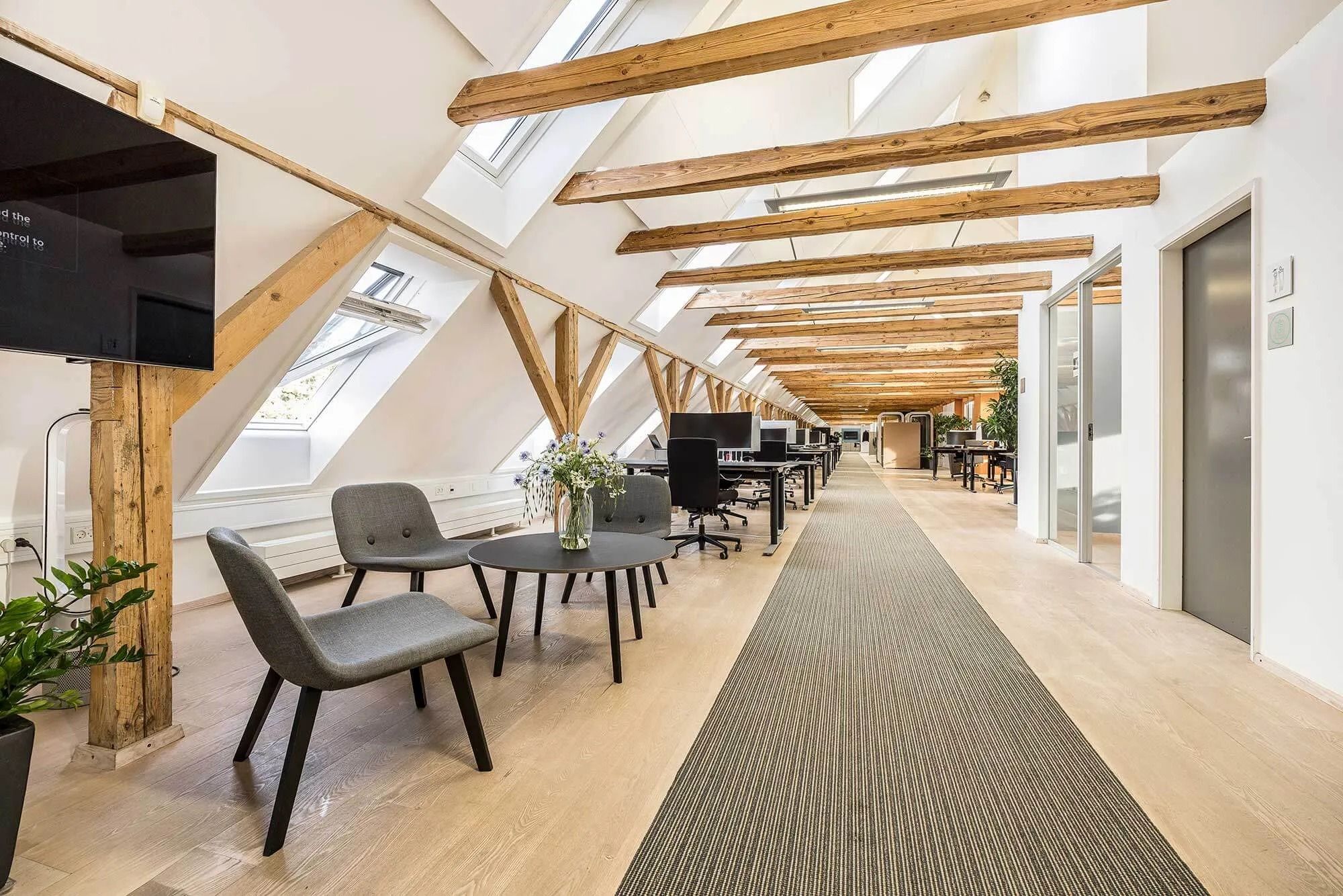 Moderne kontorlokaler med åbne lofter og lyse vinduestager, hvor der er placeret kontormøbler og et stort tv