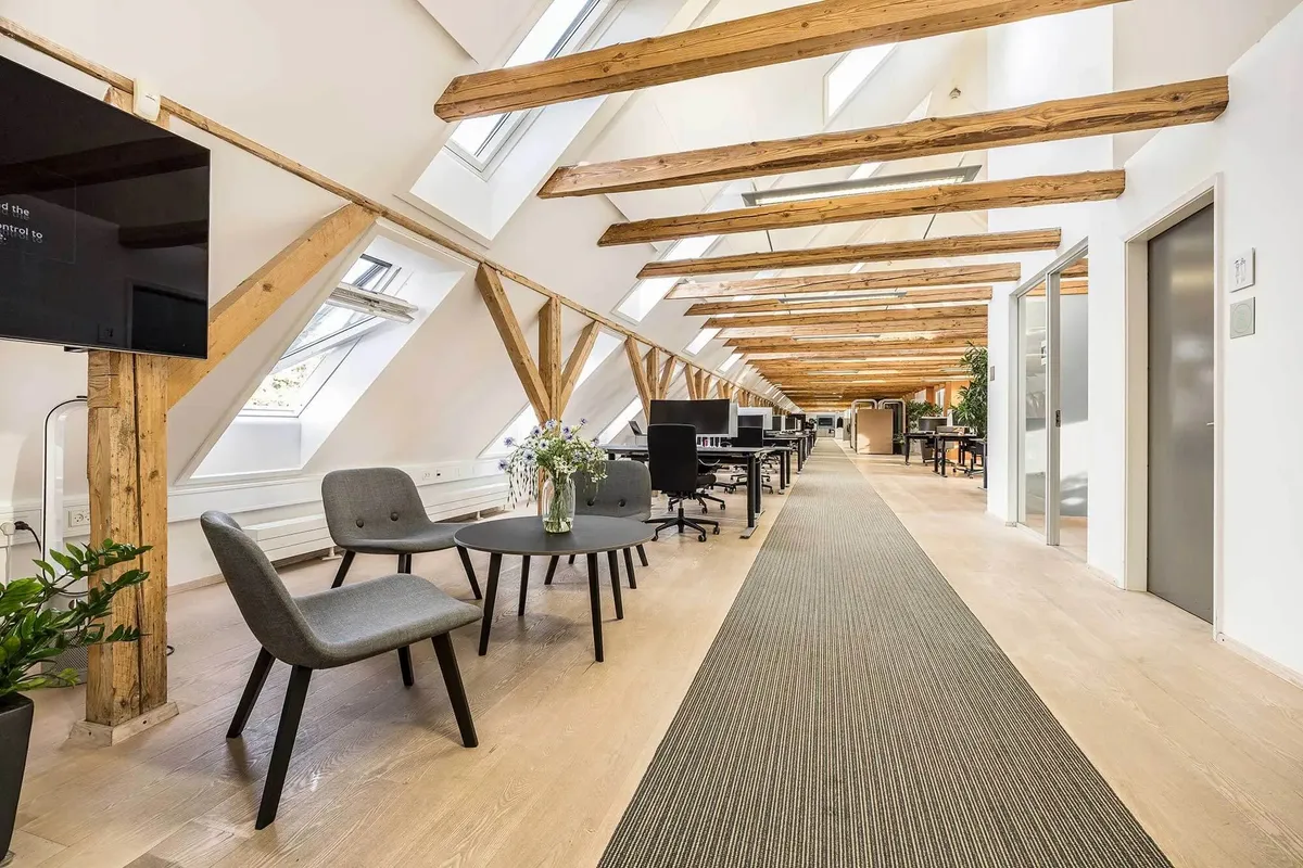 Moderne kontorlokaler med åbne lofter og lyse vinduestager, hvor der er placeret kontormøbler og et stort tv
