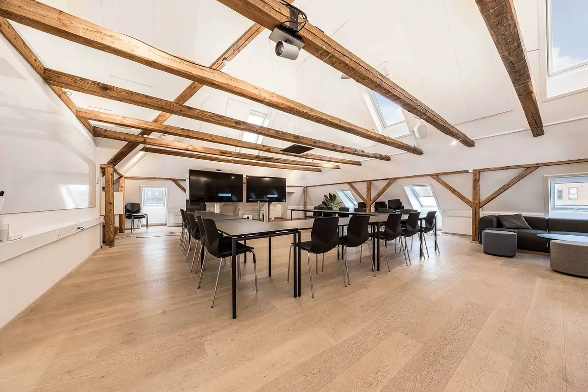 Kontorloft med lyse træbjælker og lyse loftsvinduer samt en stor mødesal med lange borde og stole