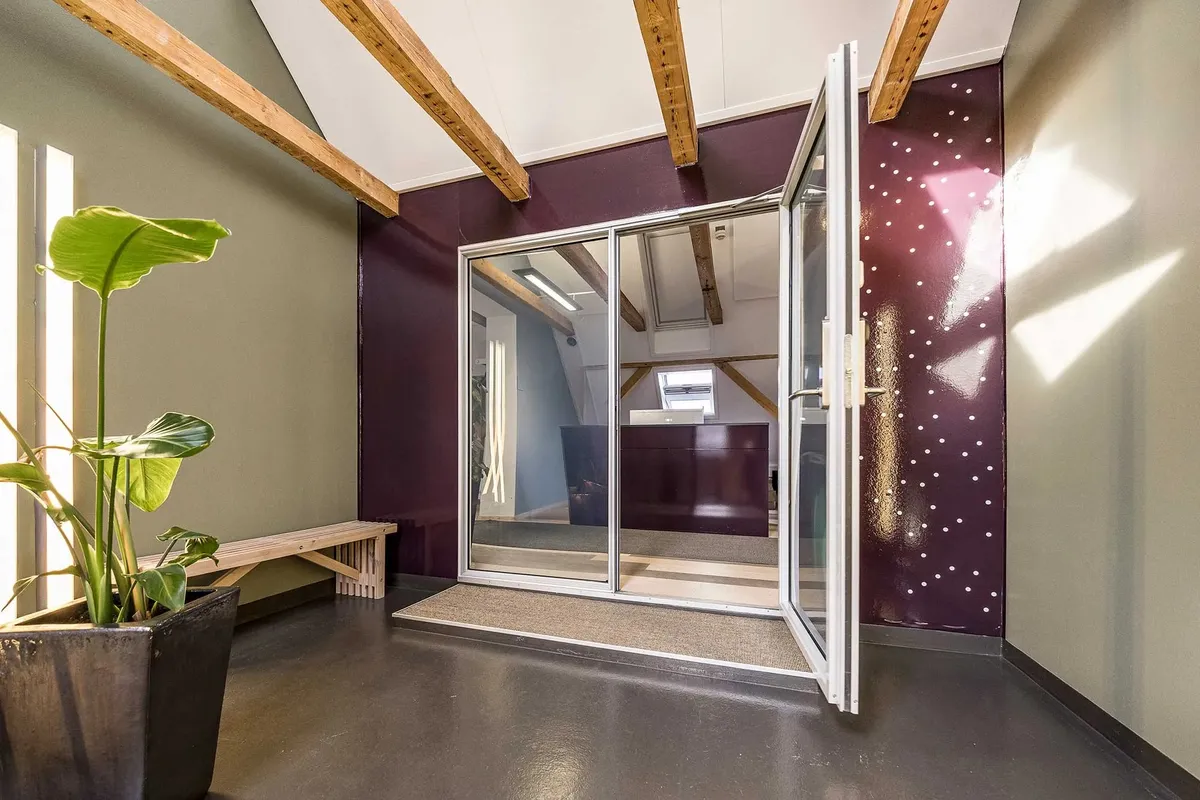 Indtrædszone til moderne kontorejendom med åben dør, beklædt med lysegrå og mørkebrune vægge, indret med møbler og plante i