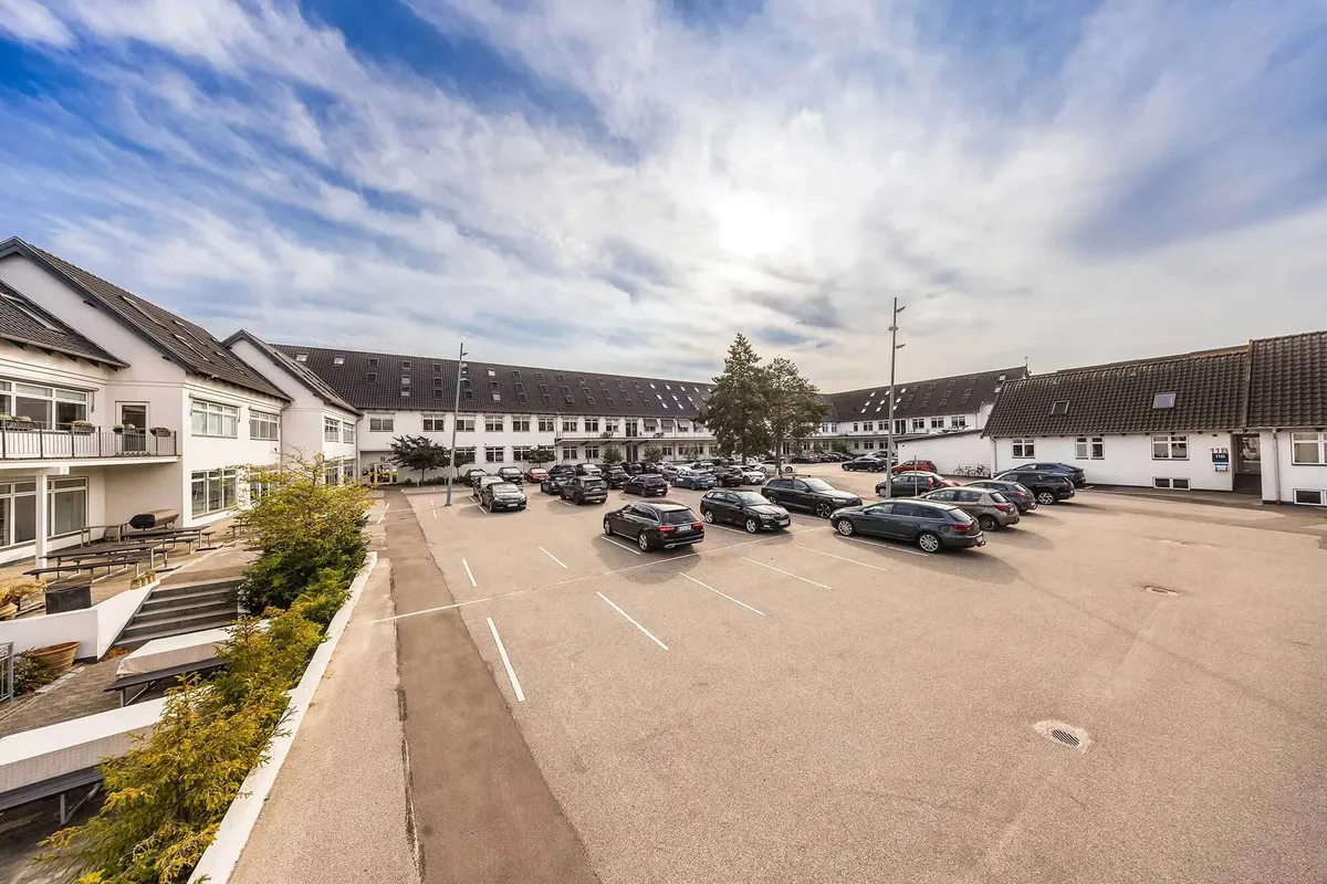 Moderne bygning med parkeringsplads og grønne planter udenfor under en delvist skyet himmel