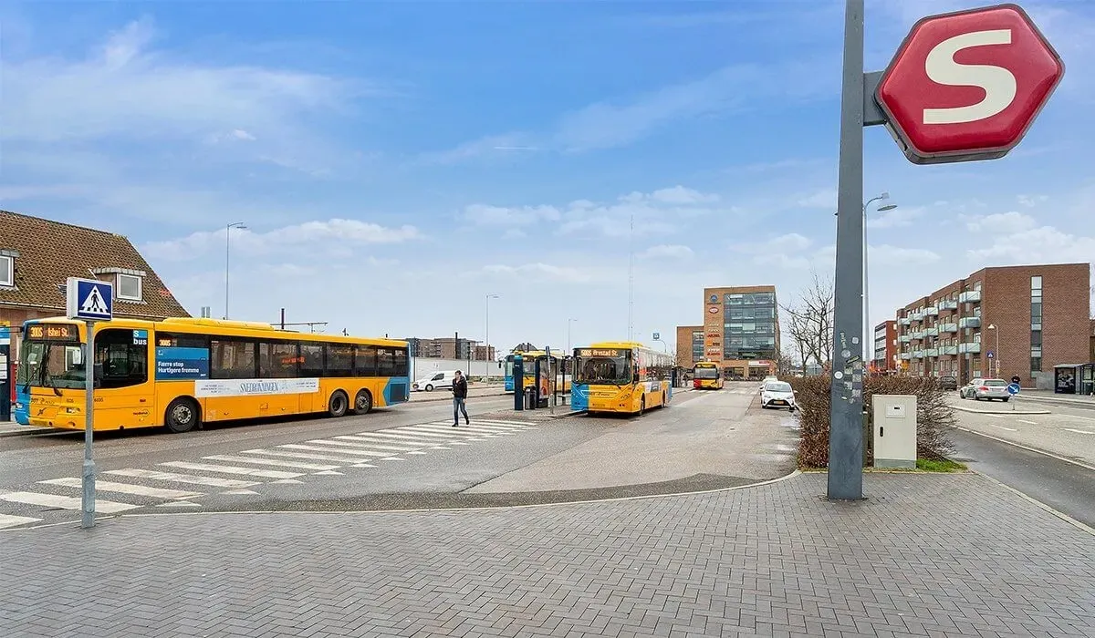 Busstation med tre guldfarvede busser og en stopskilt med bogstavet S på et sted med bebyggelse og veje