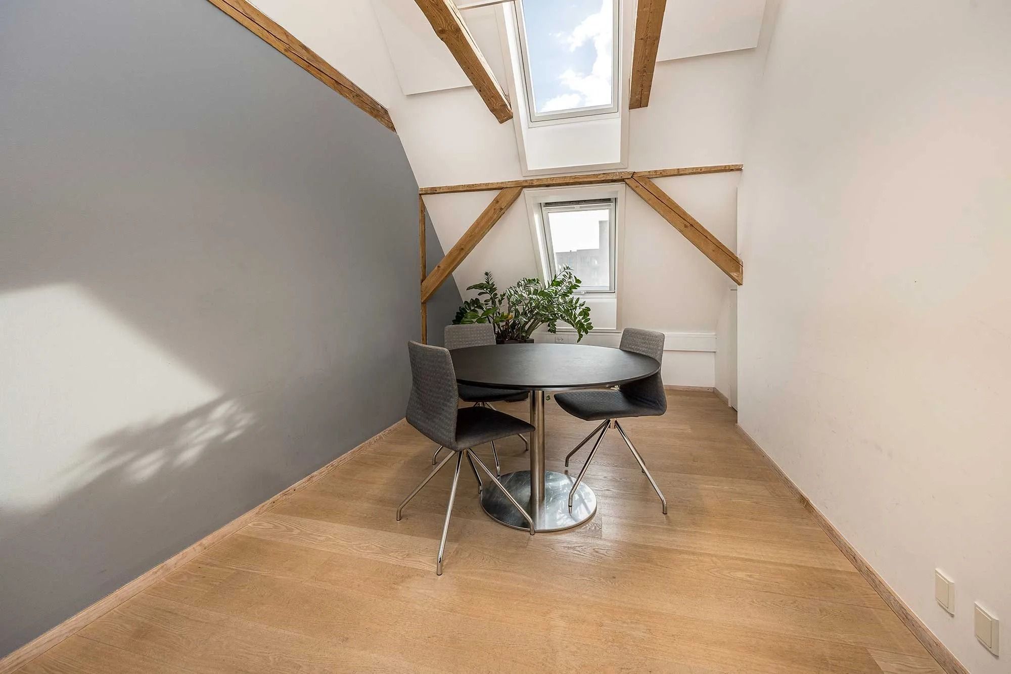 Moderne loftsværelse med træloft og lyseruder, rundt bord og stole, samt et grønt plante.