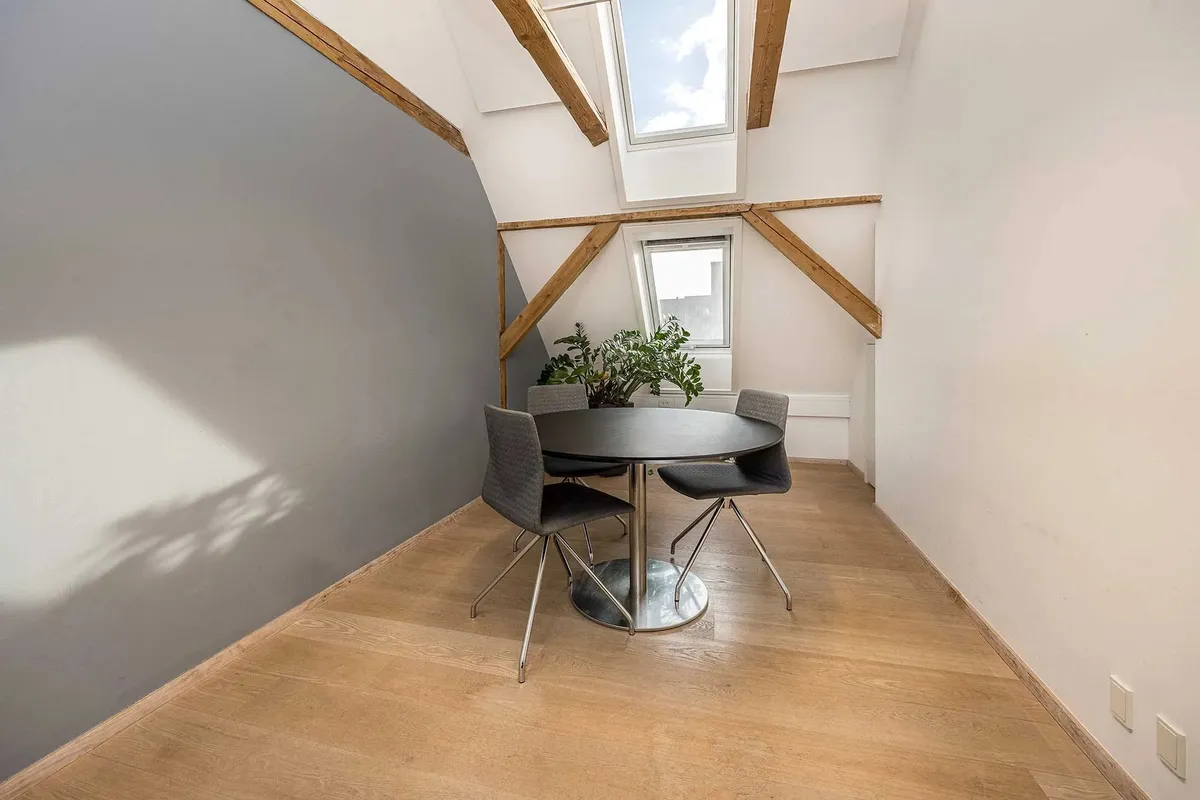 Moderne loftsværelse med træloft og lyseruder, rundt bord og stole, samt et grønt plante.