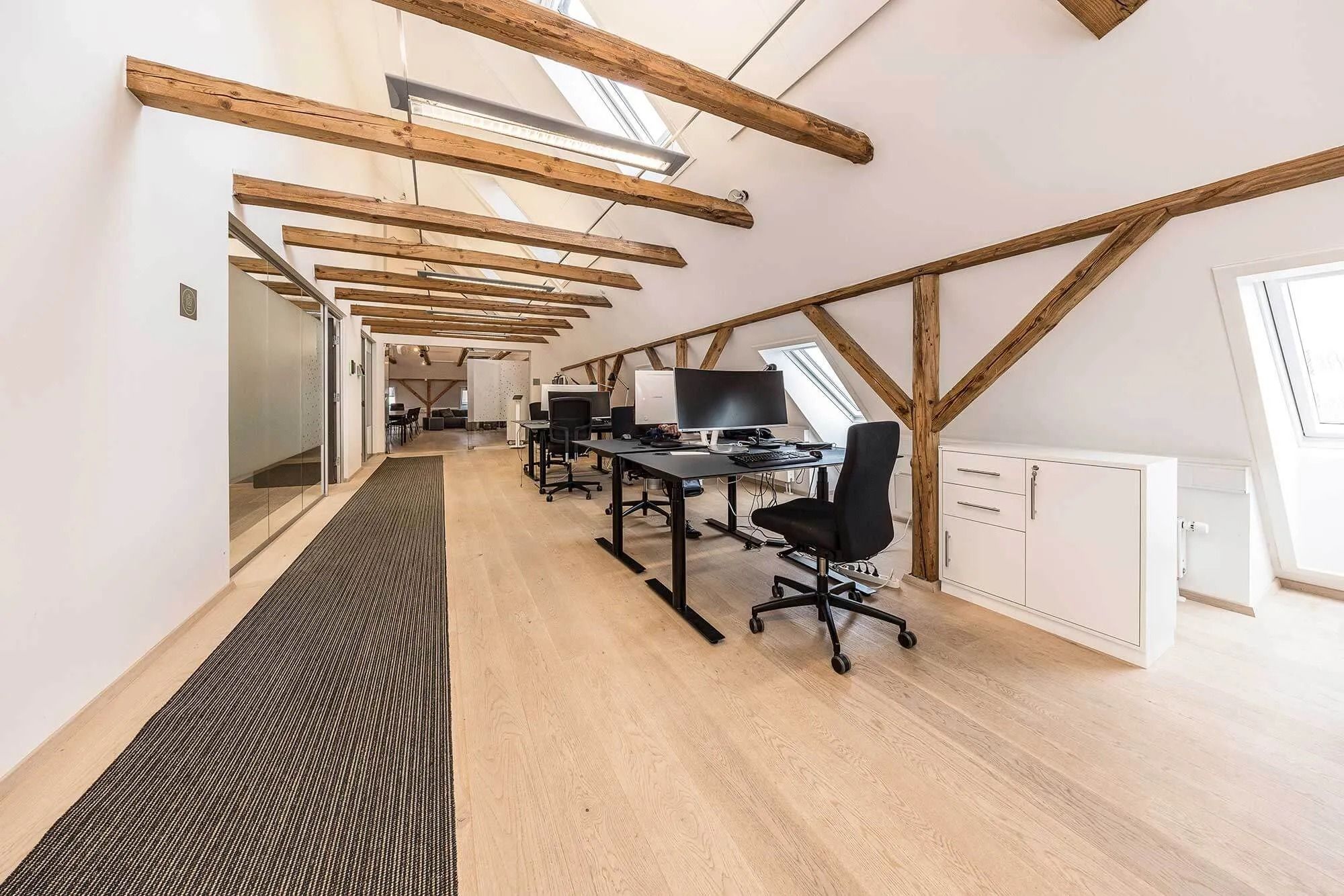 Kontorrum med moderne møbler, computere, højloft og lyse loftsbjælker i lysegrå og hvid farve