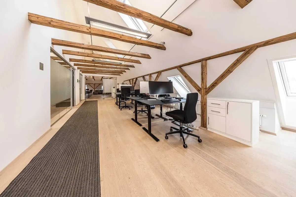 Kontorrum med moderne møbler, computere, højloft og lyse loftsbjælker i lysegrå og hvid farve
