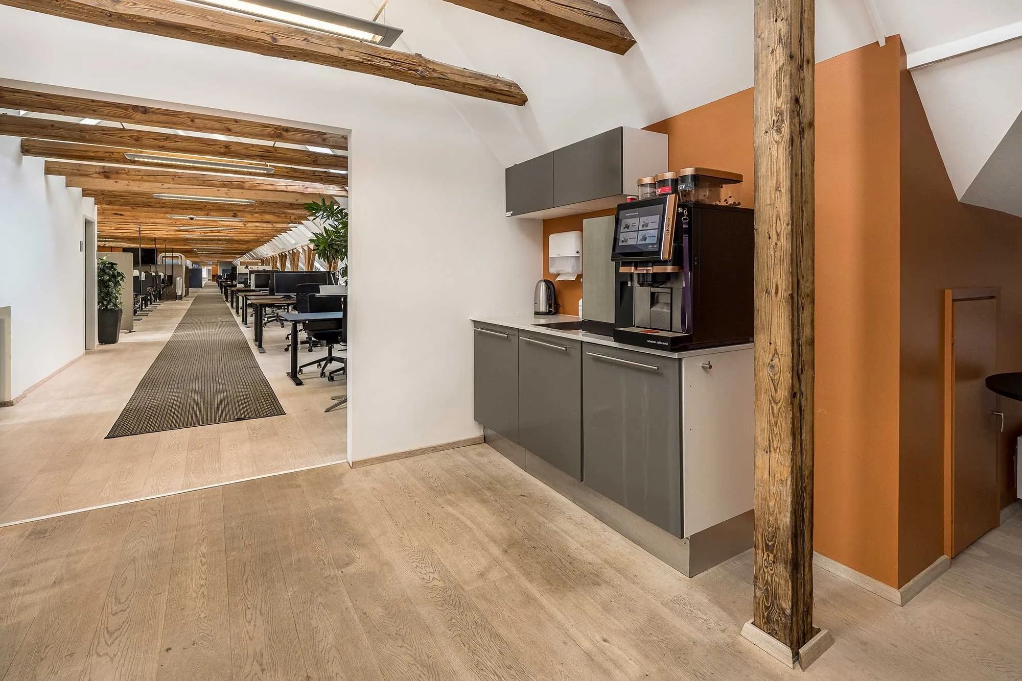 Moderne kontorområde med lyse loftsbjælke, trægulve og køkken med kaffemaskine, kedel og vask samt møbler og planter langs