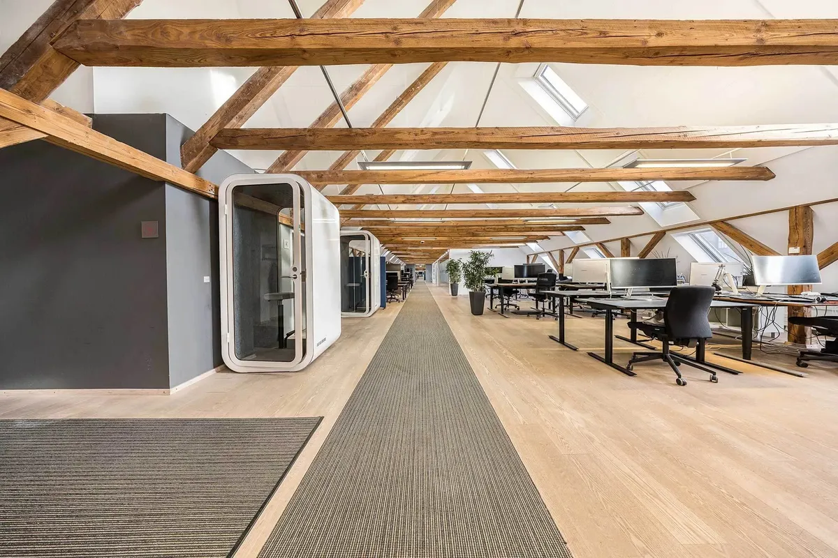 Kontor med lyse lofter og træbjælker, høje vinduer og et langt arbejdsbord med computere og stole