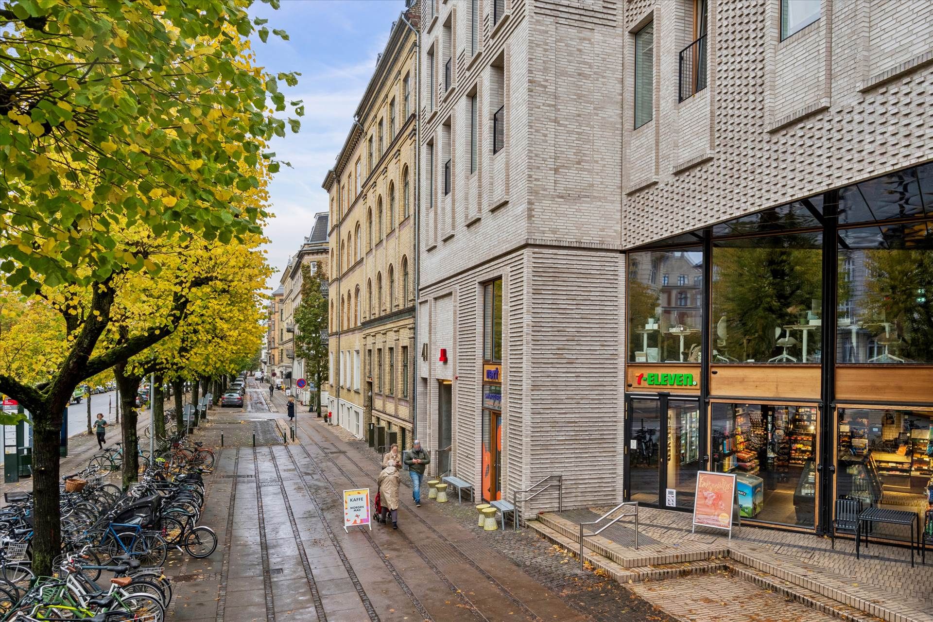 En moderne bygning med glasruder og teglvægge, hvor der står 7-Eleven. Folk går på gaden, og der er træer og cykler langs