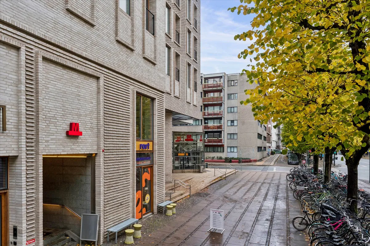 En række cykler parkeret ved siden af en stor bygning og træer med gule og grønne blade og et lysrødt bogstav M på en mur