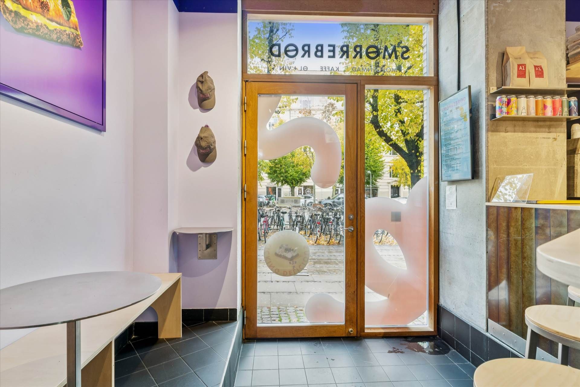 Indgangsdør til en cafe med skiltet "Verners" i rødt og gult. Indvendigt er der træborde, træstole, og et bagerehylle med