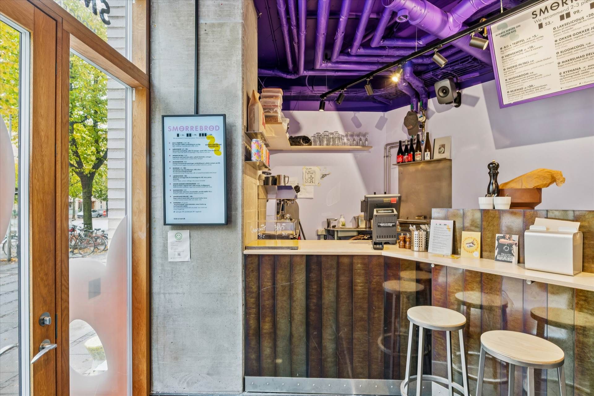 Et café med mørke vægge og lyse gulve, en menu på væggen, flere høje stole, og flere glas og flasker på en hylde