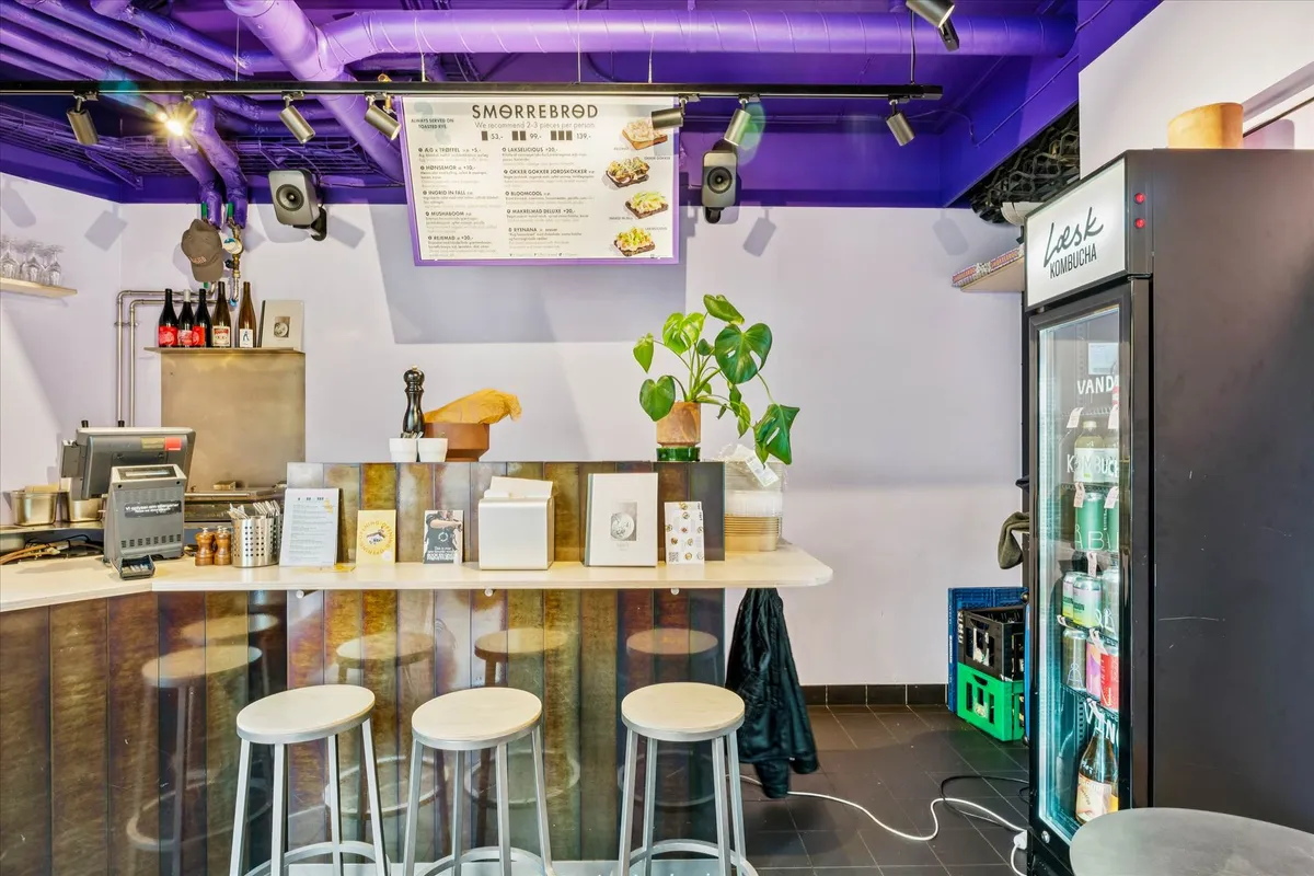 Køkken med højt barbord, tre høje stole, menutavle, komfur, skab med glasdøre, blomster og drikkevarer i køleskab samt