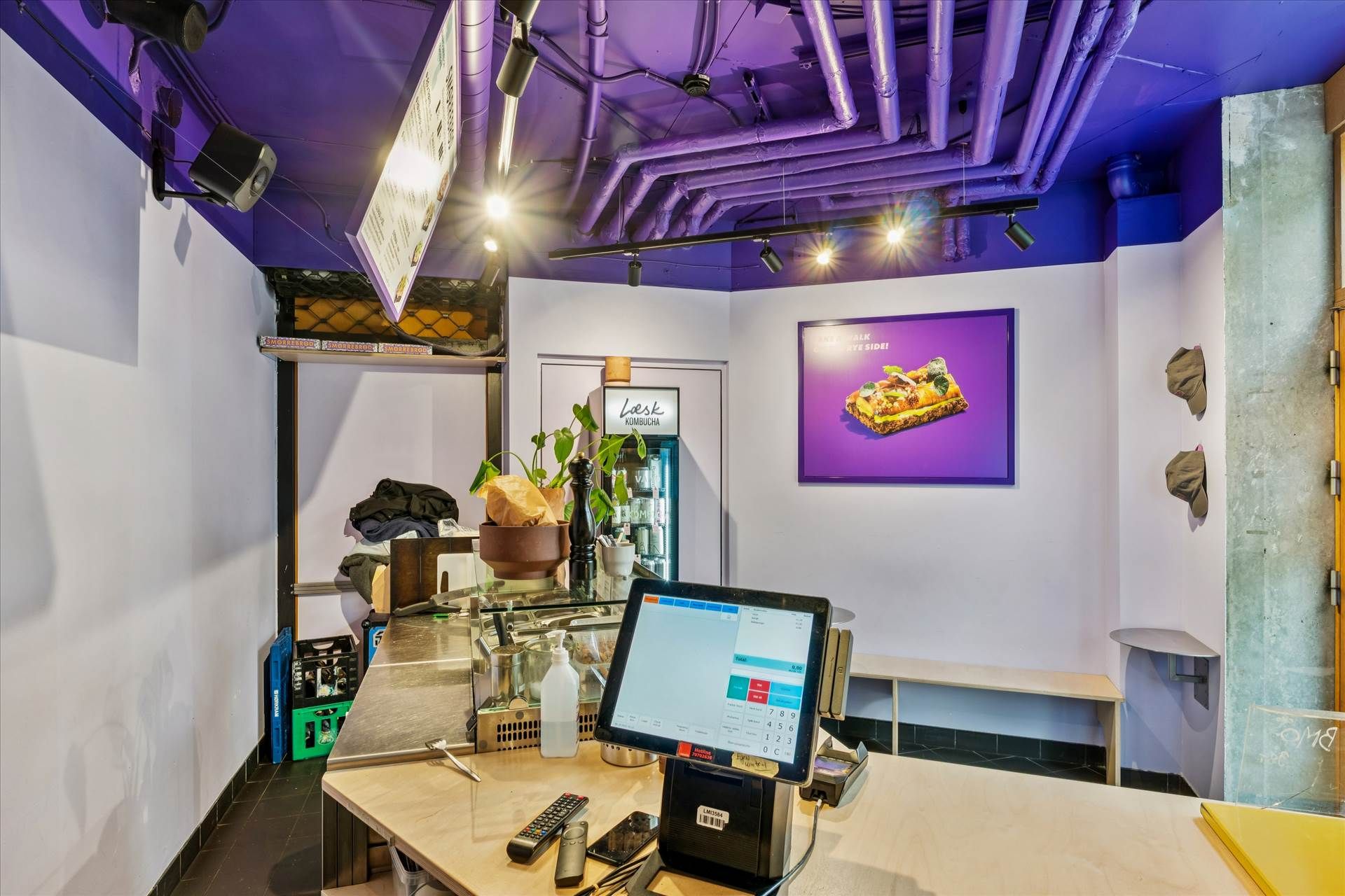 En sælgerkontor med en kasseapparat, fjernbetjeninger og en beholder med drikkevarer. Der er også en plante og en skilt med