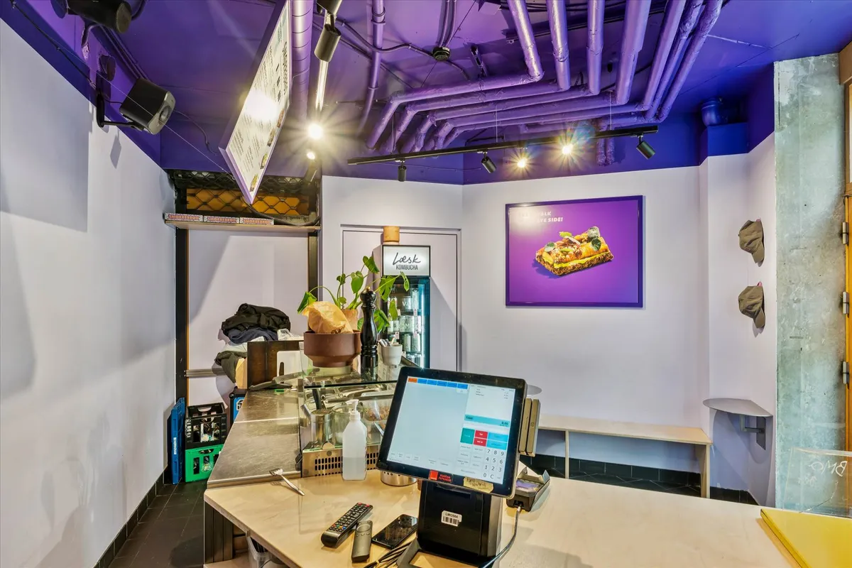 En sælgerkontor med en kasseapparat, fjernbetjeninger og en beholder med drikkevarer. Der er også en plante og en skilt med