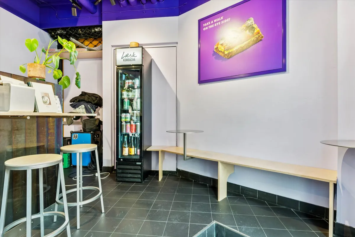 Inde i et komfortabelt lokale med moderne møbler og træelementer, hvor en kombucha-køleskab og to stolpe med høje ben er