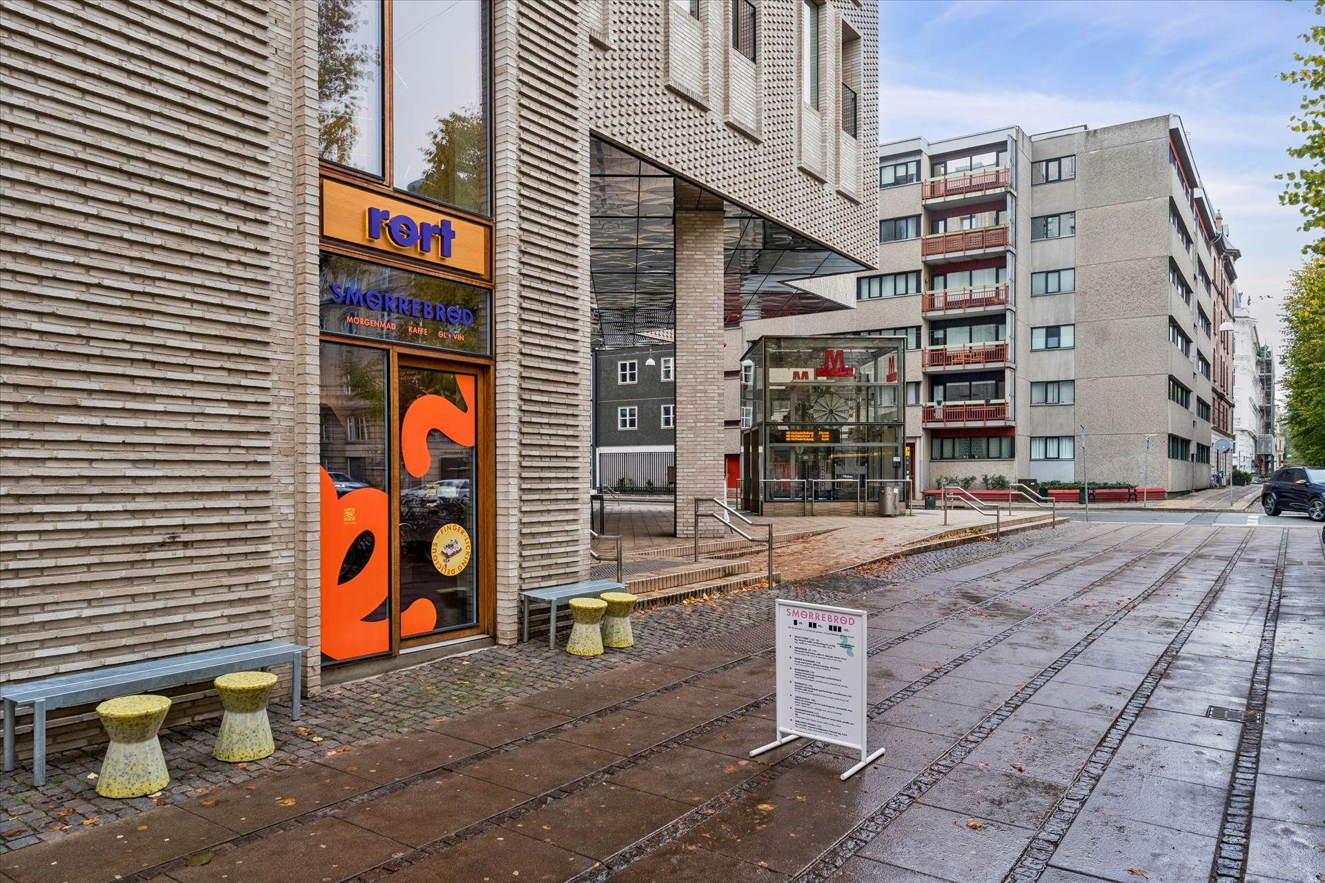 Røgfri Smorrebrød-forretning med solrige ruder og orange indgangsdør samt guldfarvede stole og bord på forsiden af