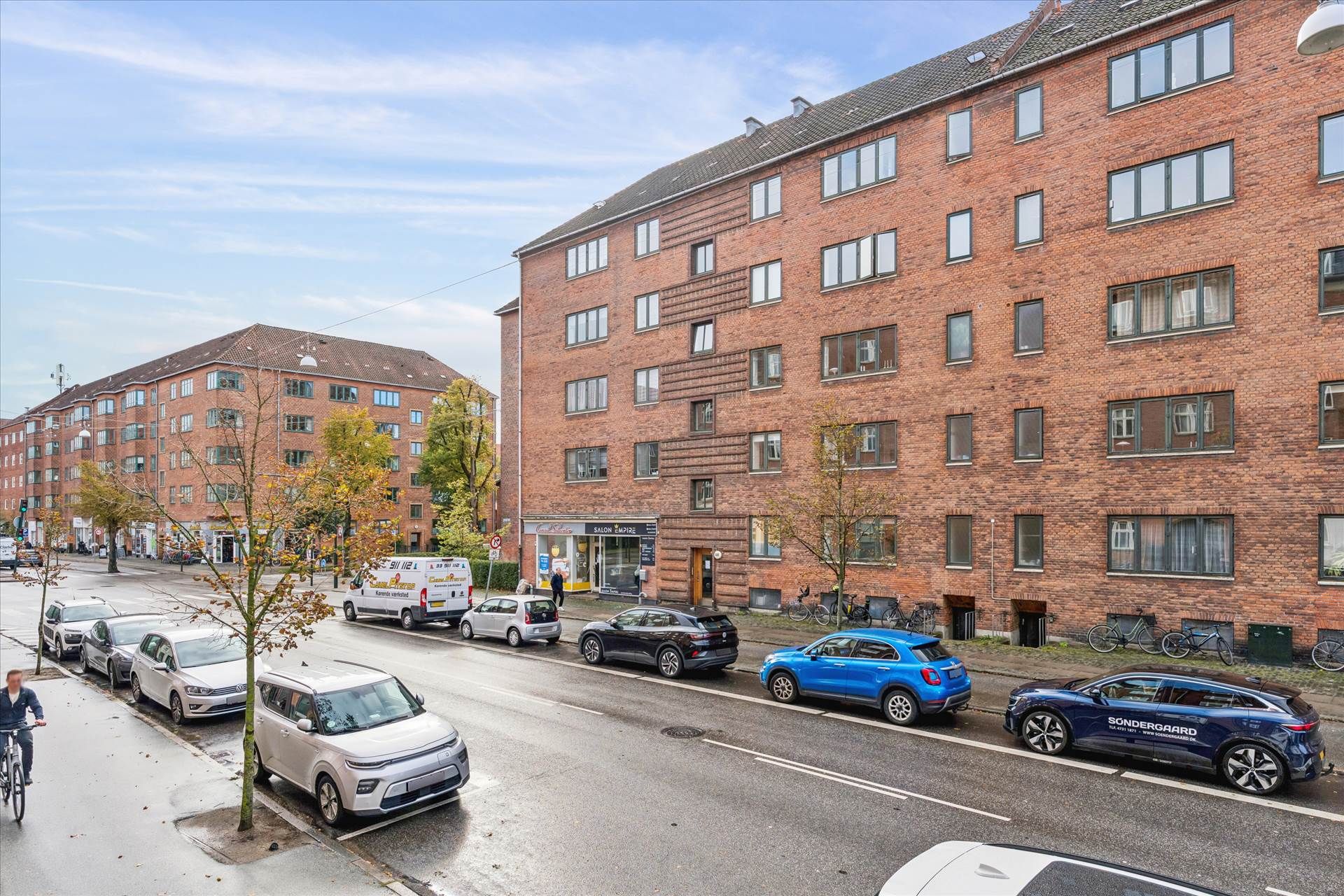 Adgangsvej med parkerede biler og mennesker i en by. Mange biler er parkeret langs siderne, og der er også en cykel.