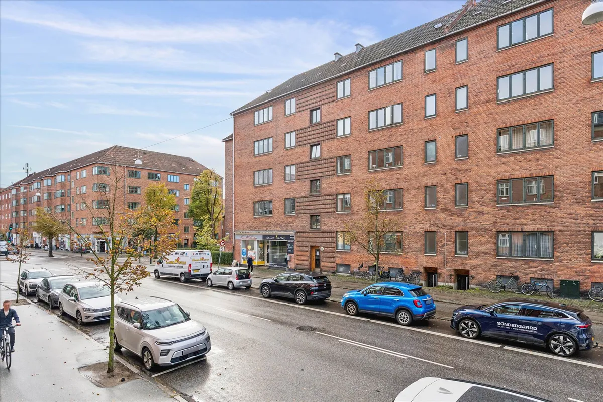 Adgangsvej med parkerede biler og mennesker i en by. Mange biler er parkeret langs siderne, og der er også en cykel.