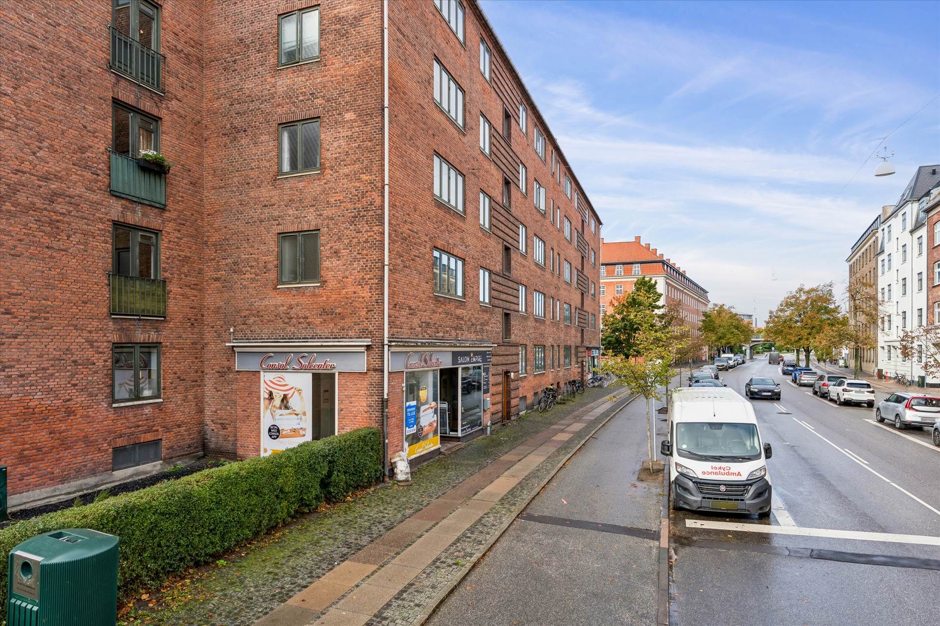 Et erhvervslejemål med flere butikker og en hvid varevogn parkeret på siden af en asfaltvej med grønne buske og træer.