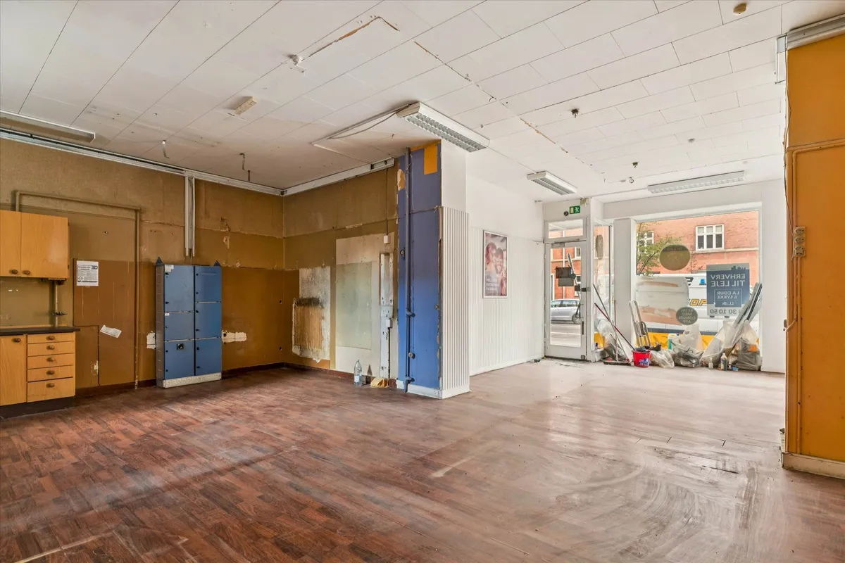 Et tomt rum med hårdt trægulv, lysegule vægge og åbne døre med lyseblå søjler samt et sammensat skriftstykke udenfor