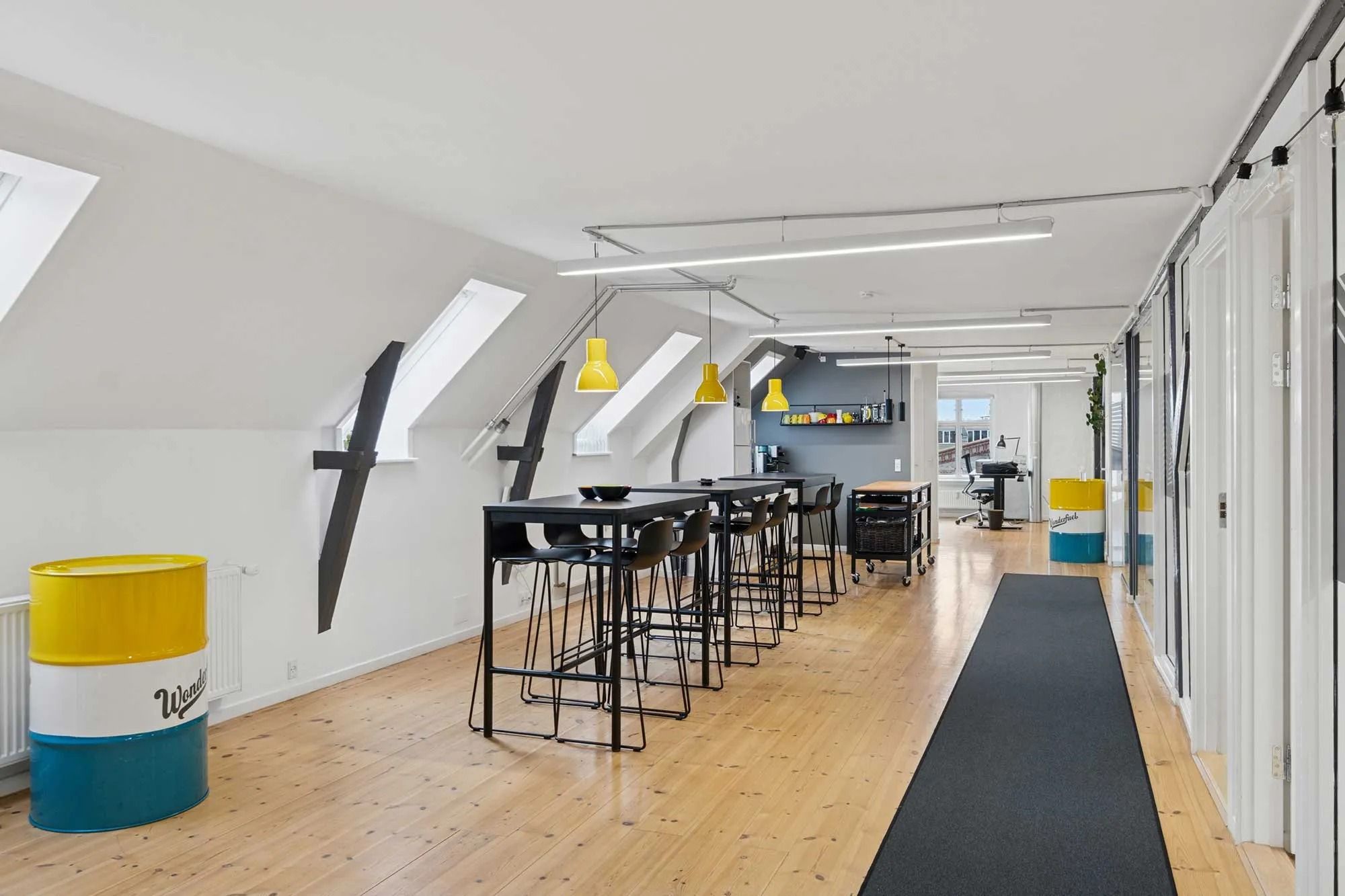 Kontorlokaler med åbne rum, højloft og lyse loftsvindue. Bord og stole samt loftsleje. Styrketøj og skraldspande i farverige