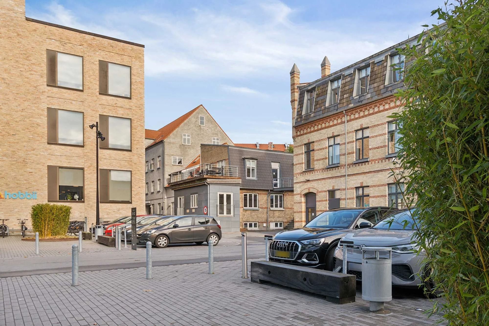 Forskellige biler parkeret udenfor et bygningskompleks med teksten Hobbit og to bænke samt en skraldespand