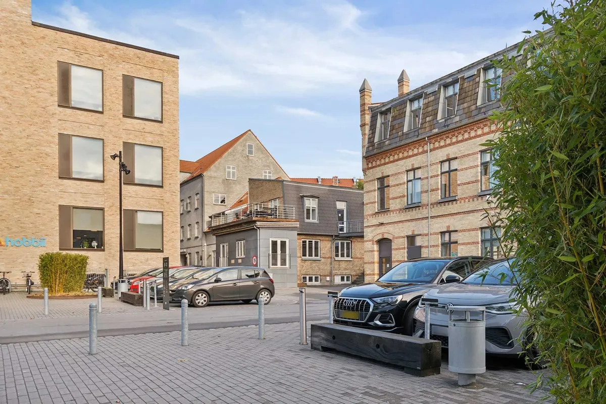 Forskellige biler parkeret udenfor et bygningskompleks med teksten Hobbit og to bænke samt en skraldespand