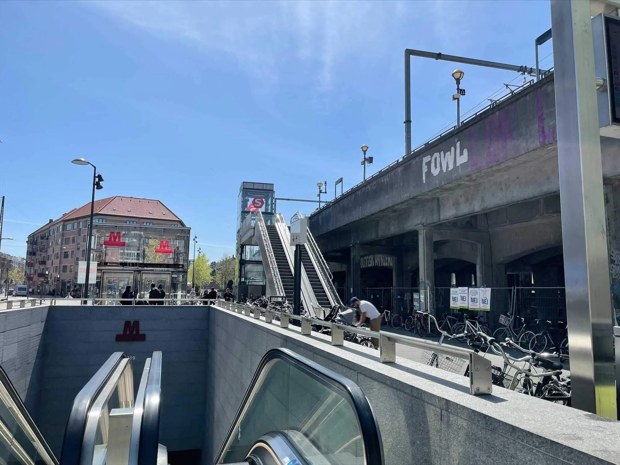 Gangbro over gade med op- og nedgangsrulletrapper, folk går på broen og en mand smider cykler fra trappen
