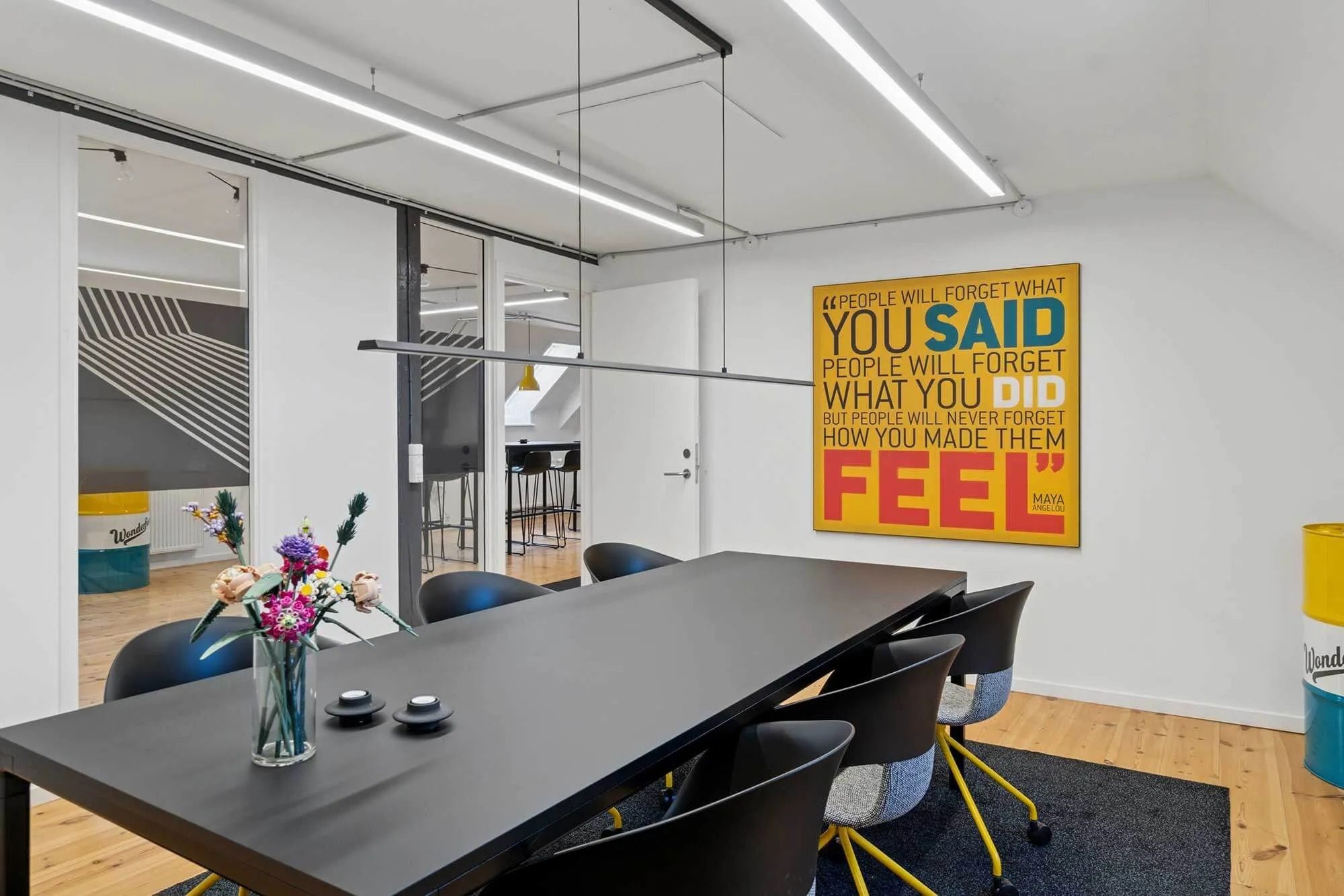 Kontorrum med lyse loftslampe, møbler og kunstværk på væggen. Der er en stor bord og stol i midten af rummet.