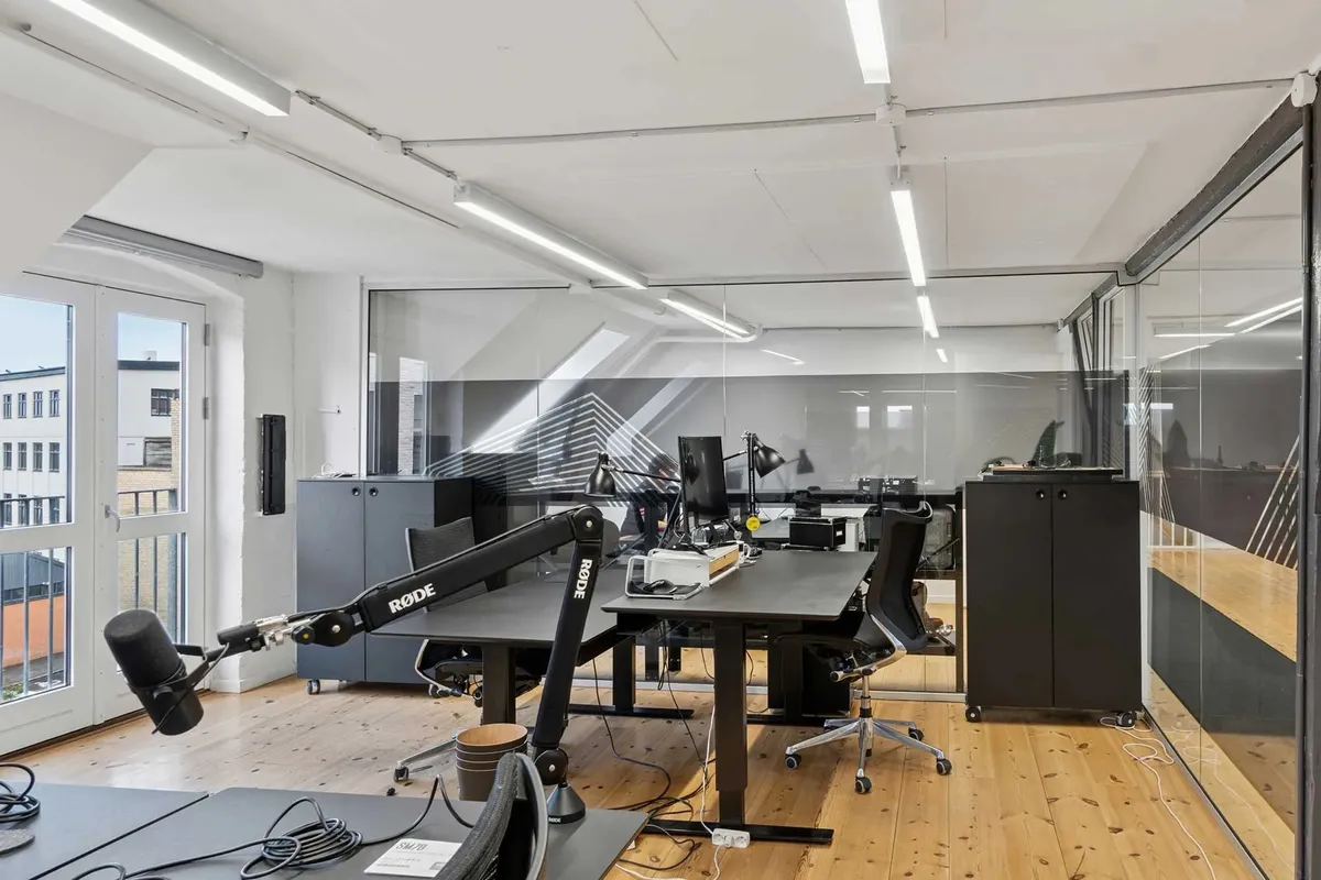 Kontor med flere bordsæt og skriveborde, højteknologisk udstyr, flere computere, kabinetter og vinduer med udsigt til gaden