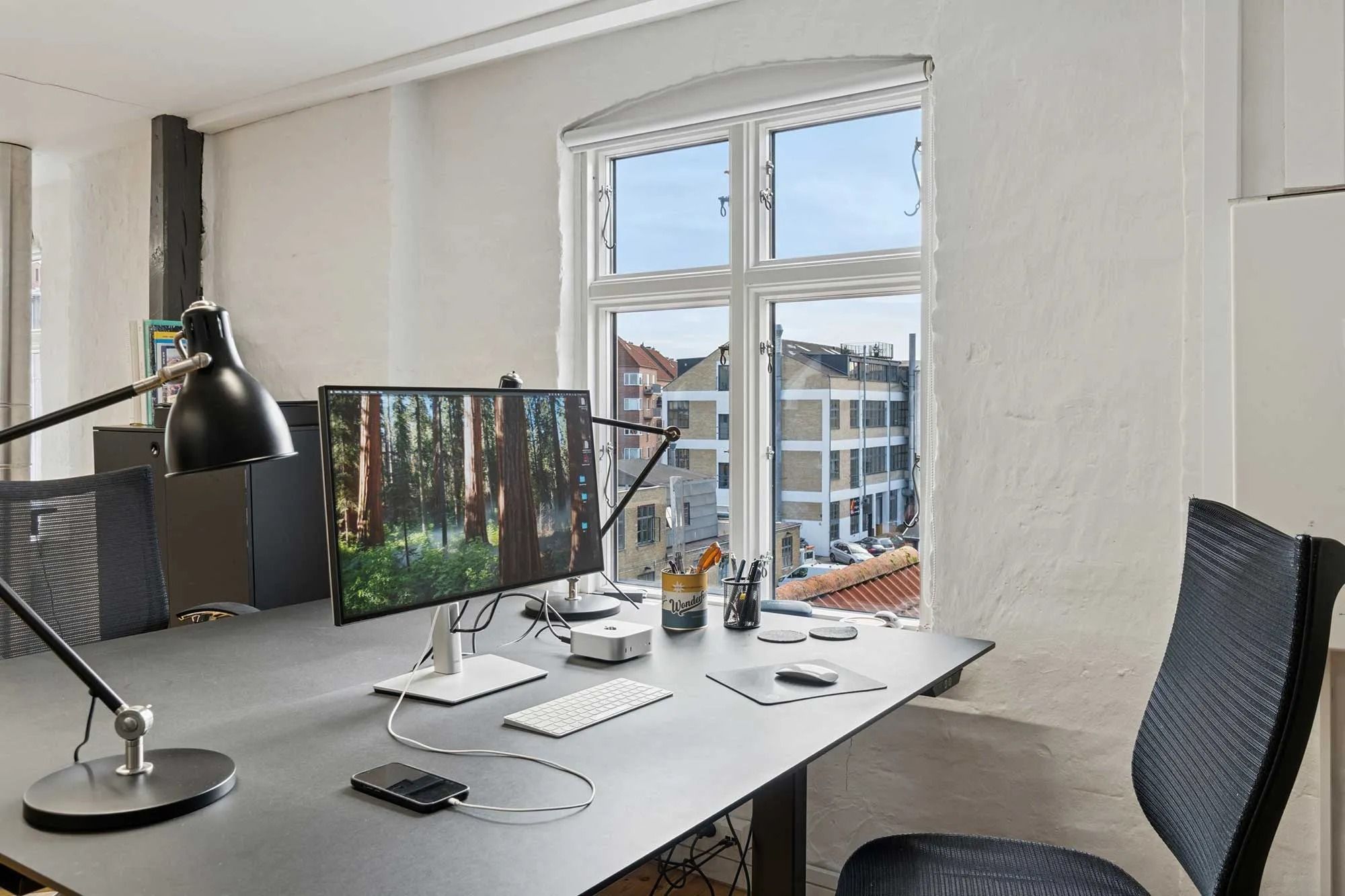 Kontor med moderne indretning, to skærme, skrivebord, skrivebordslampe, pen og blyant stående i vandglas, mobiltelefon og