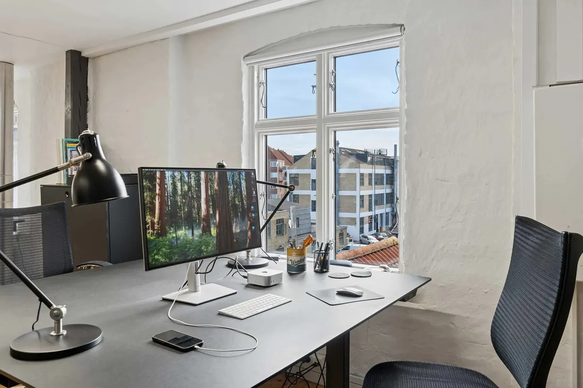 Kontor med moderne indretning, to skærme, skrivebord, skrivebordslampe, pen og blyant stående i vandglas, mobiltelefon og