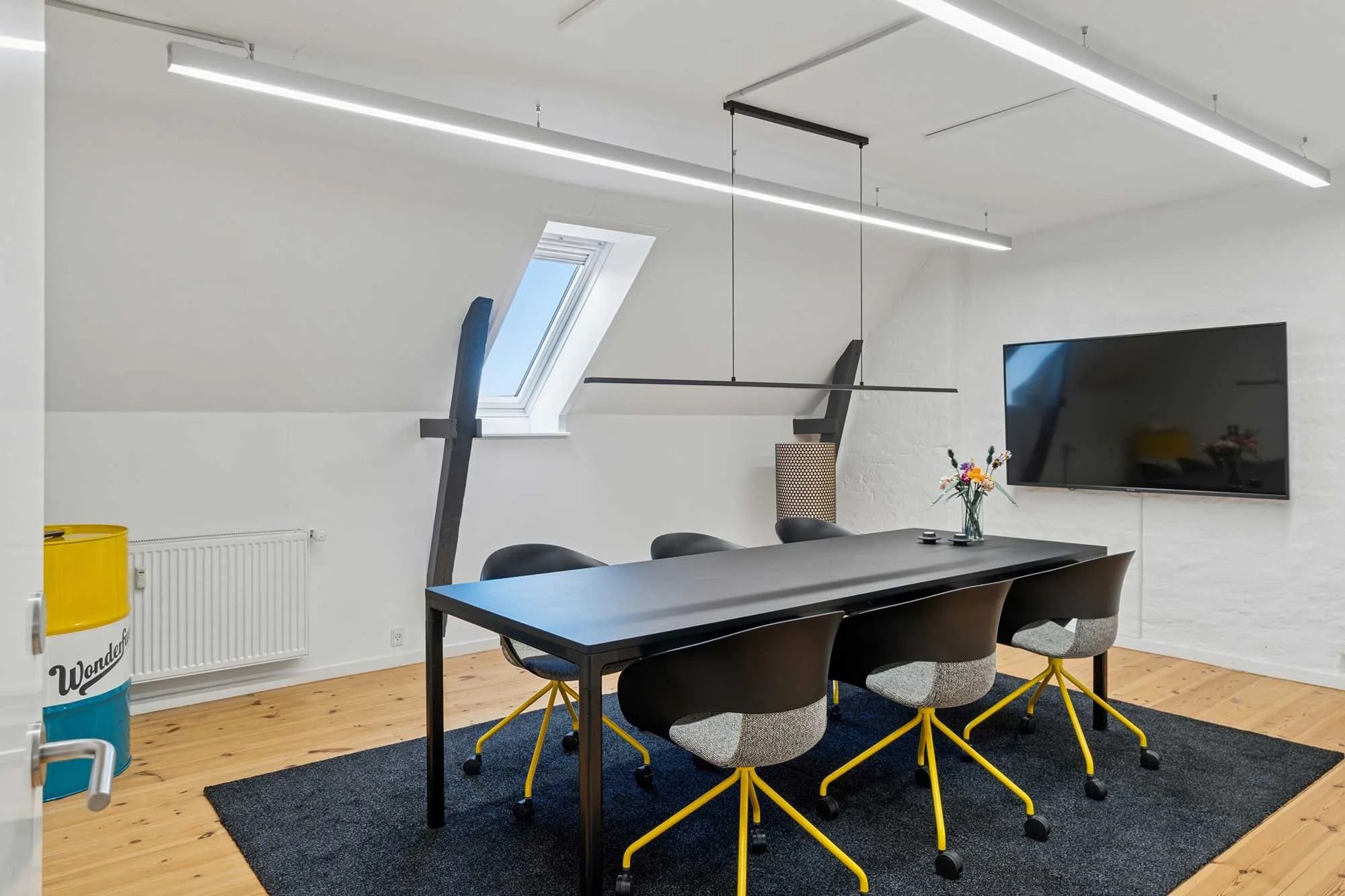 Kontor med langt bord og stålstole med hjul, tv og lysekroner samt vindue i loftet og gulvvarmer ved væggen