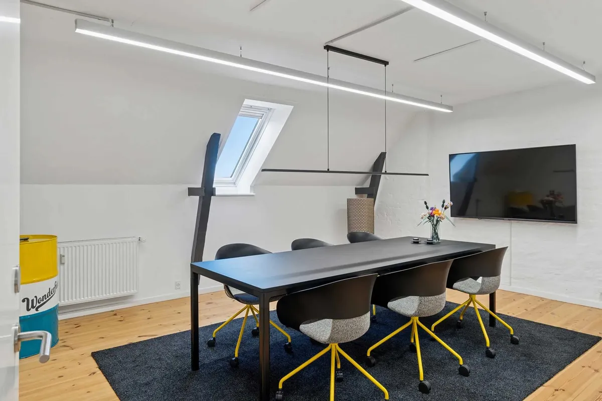 Kontor med langt bord og stålstole med hjul, tv og lysekroner samt vindue i loftet og gulvvarmer ved væggen