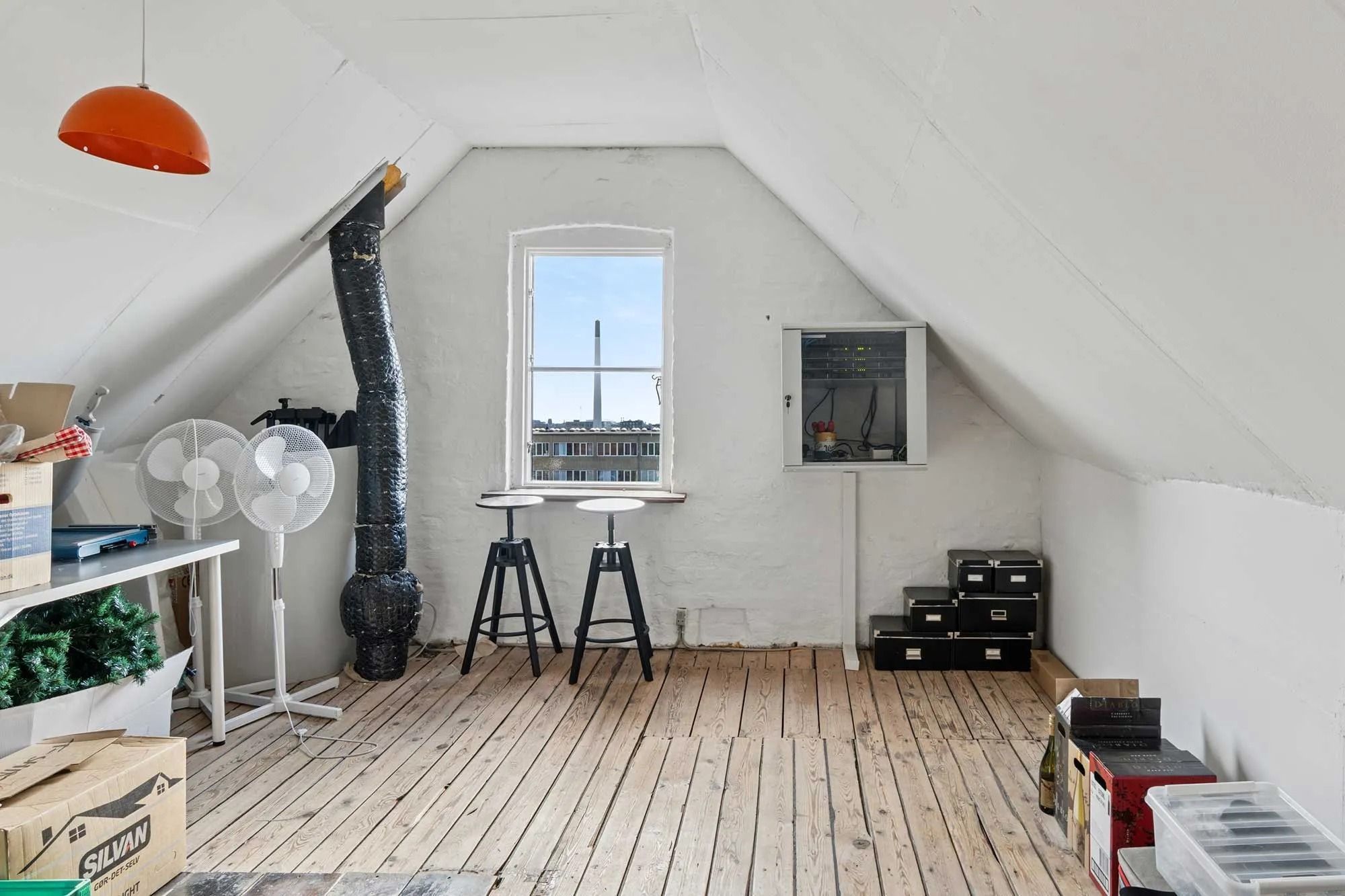 Loftsværelse med vinduer og trægulv, hvor der står et bord, stole, faner og lagerkasser med ting