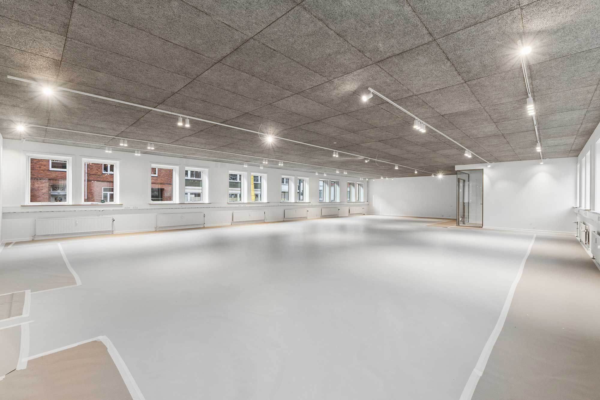 Et stort, lyse, ubenyttet rum med hvide vægge og gulv samt flere vinduer og lysekroner