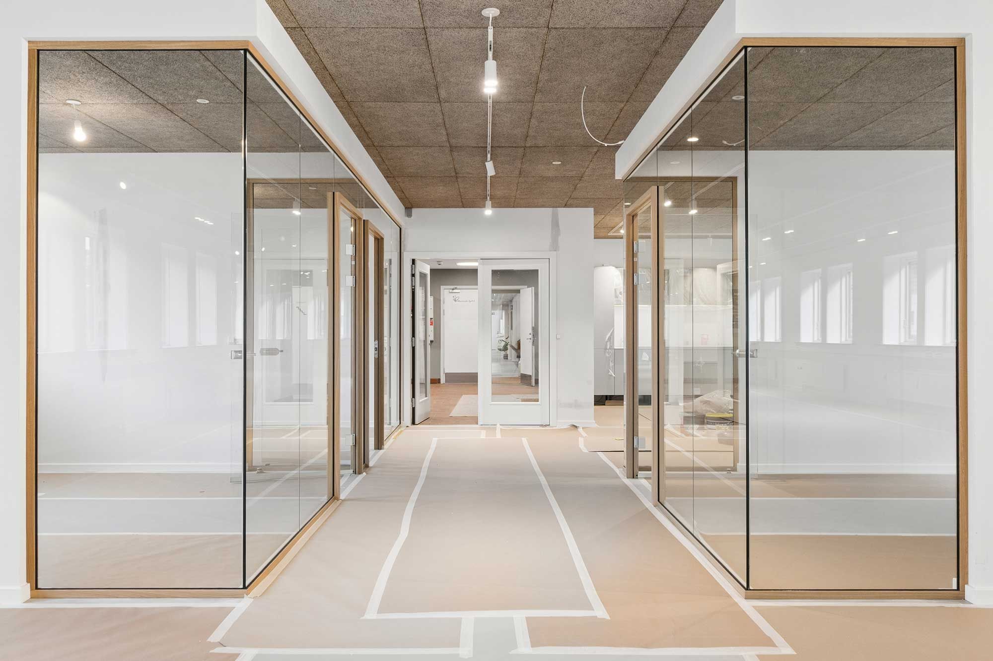 Kort og ren gange med glasvægge og lyse loftsbelægning samt åbne døre med håndtag og søjler i lysebrunt træ
