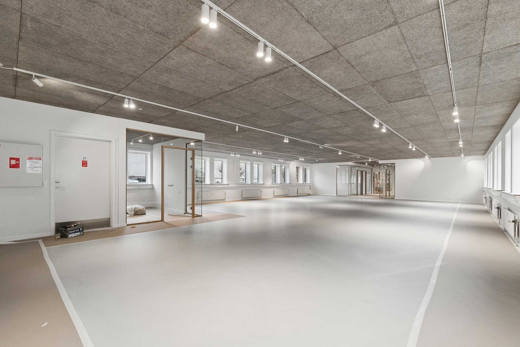 Stort værelse med hvidt loft og gulv, lysekroner og døre. En stol og et sæt værktøj står på gulvet. Parkeringsplads med flere vinduer og døre i baggrunden