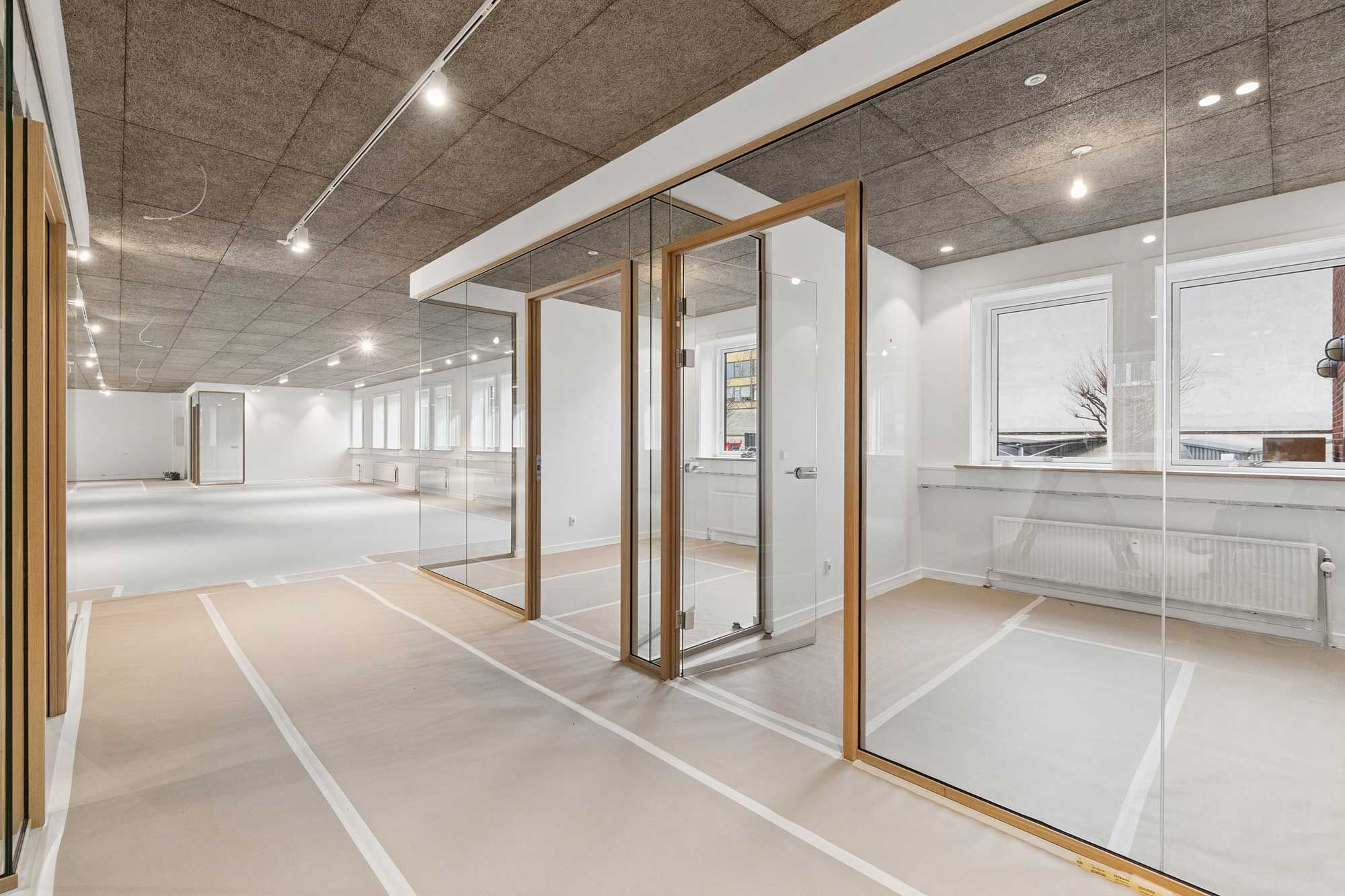 Et rummeligt og moderne kontor med glasvægge og store vinduer, trægulve, hvide vægge og et nyt centralt ventilationssystem