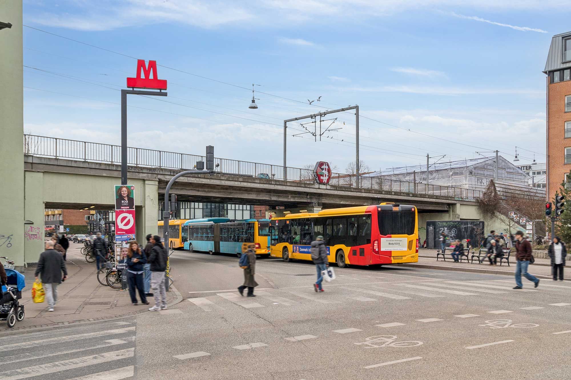 To busser står i krydset og mennesker krydser vejen, et stort M i rødt på en lysstolpe og en stor bygning i baggrunden