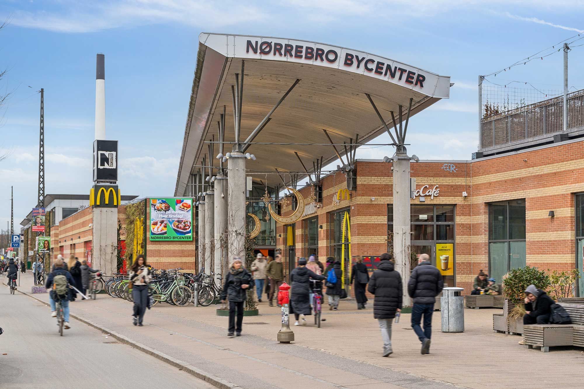 Centralt beliggende shoppingcenter med McDonald's og Café. Mange mennesker og cykler langs gågaden med udgang til S-tog og metro