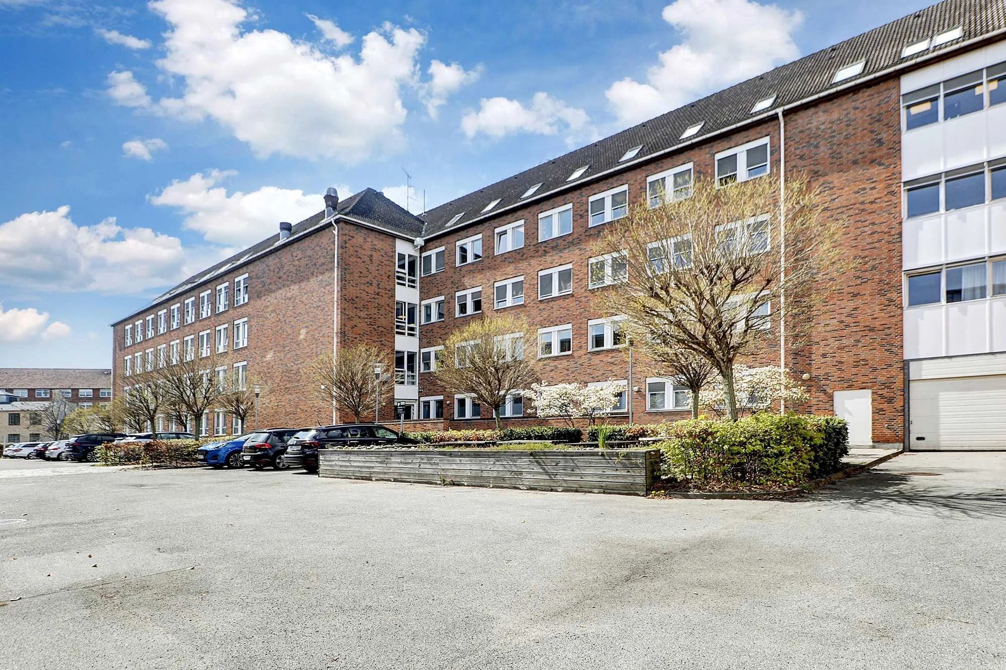 P-plads med flere biler parkeret, og buske og træer foran. Moderne bygning med røde mursten og flere vinduer.