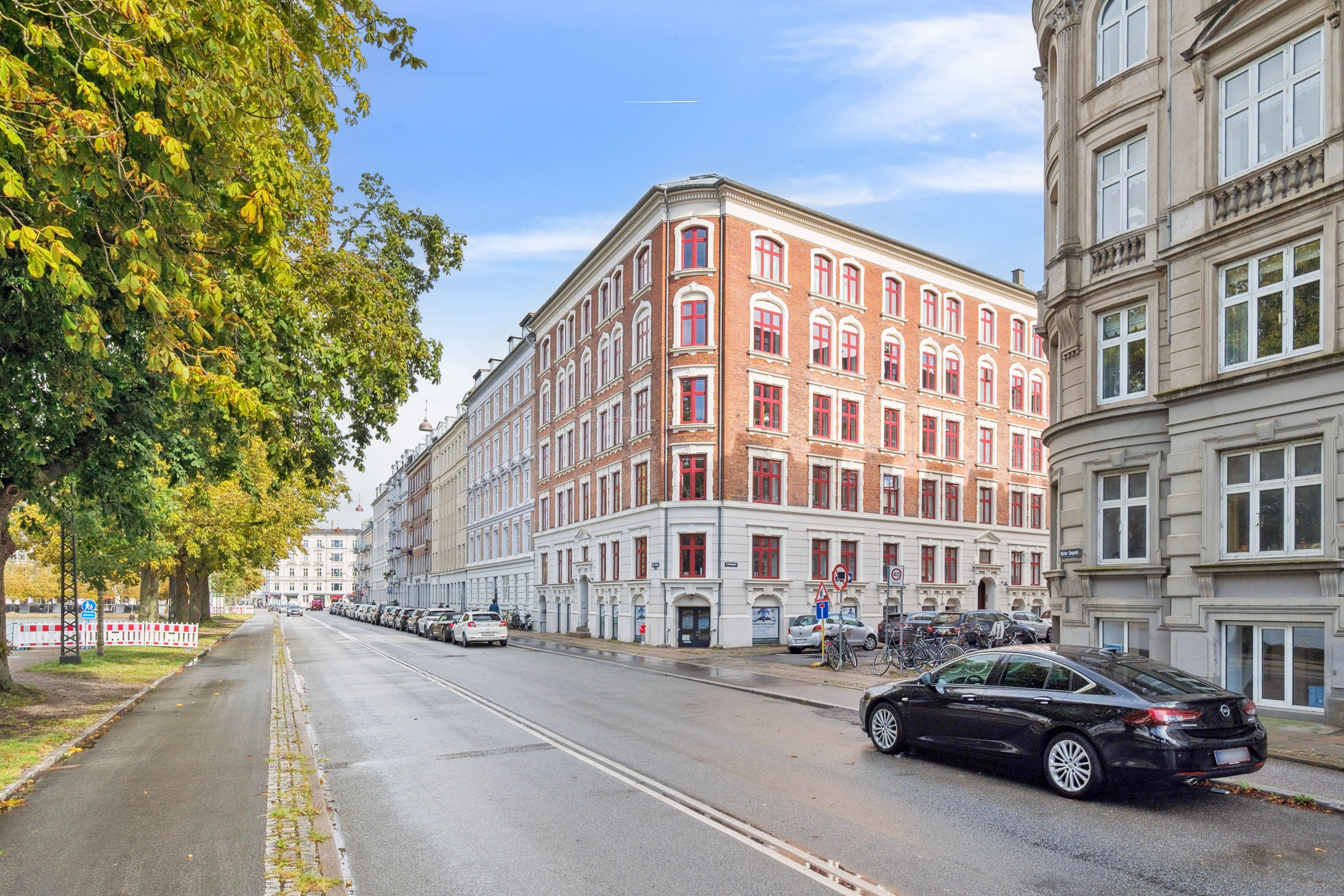 En bil kører langs en gade med røde vinduede bygninger, der vender ud mod gågaden med høje træer