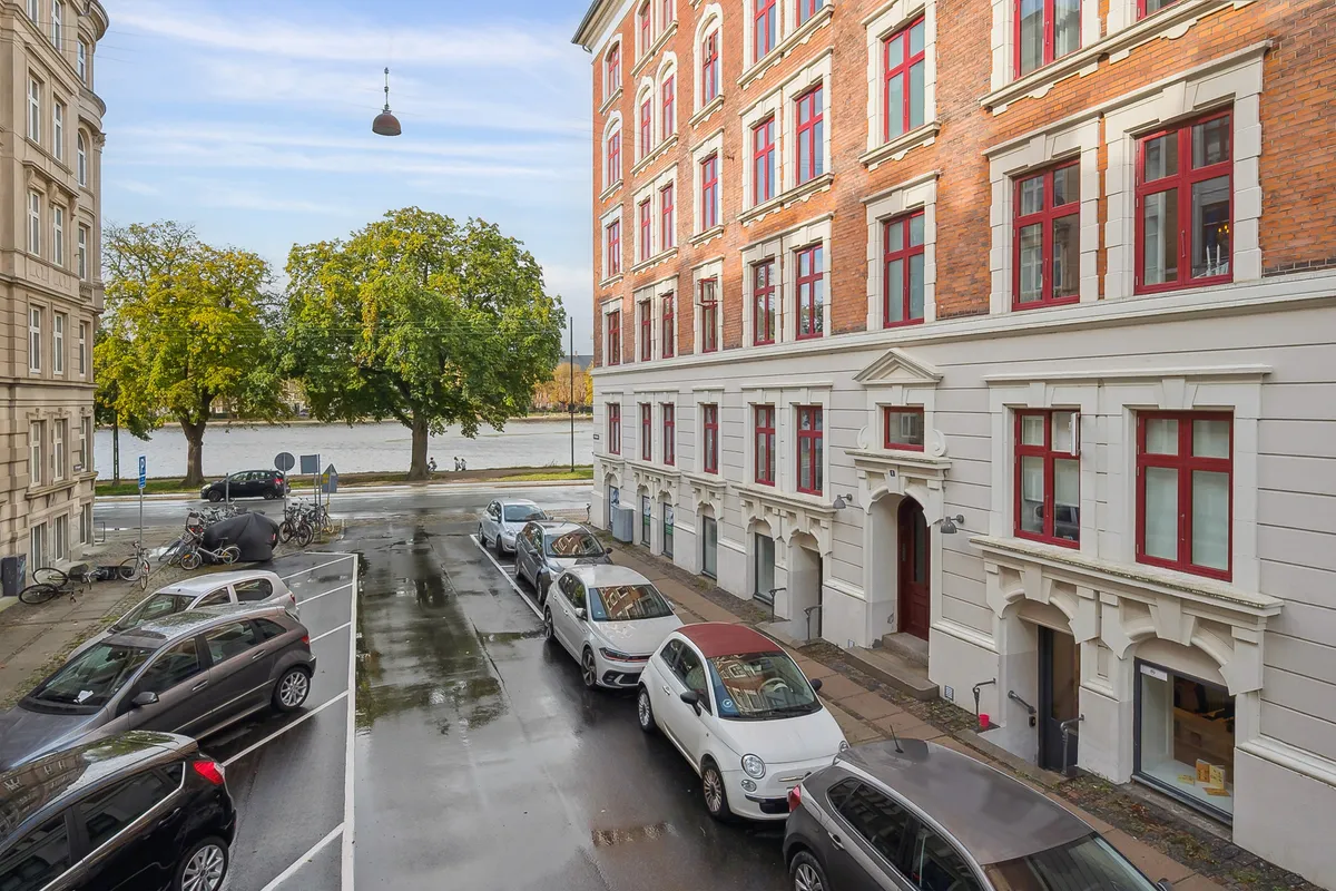 Flere biler parkeret langs en gågade med en stor bygning og sø i baggrunden