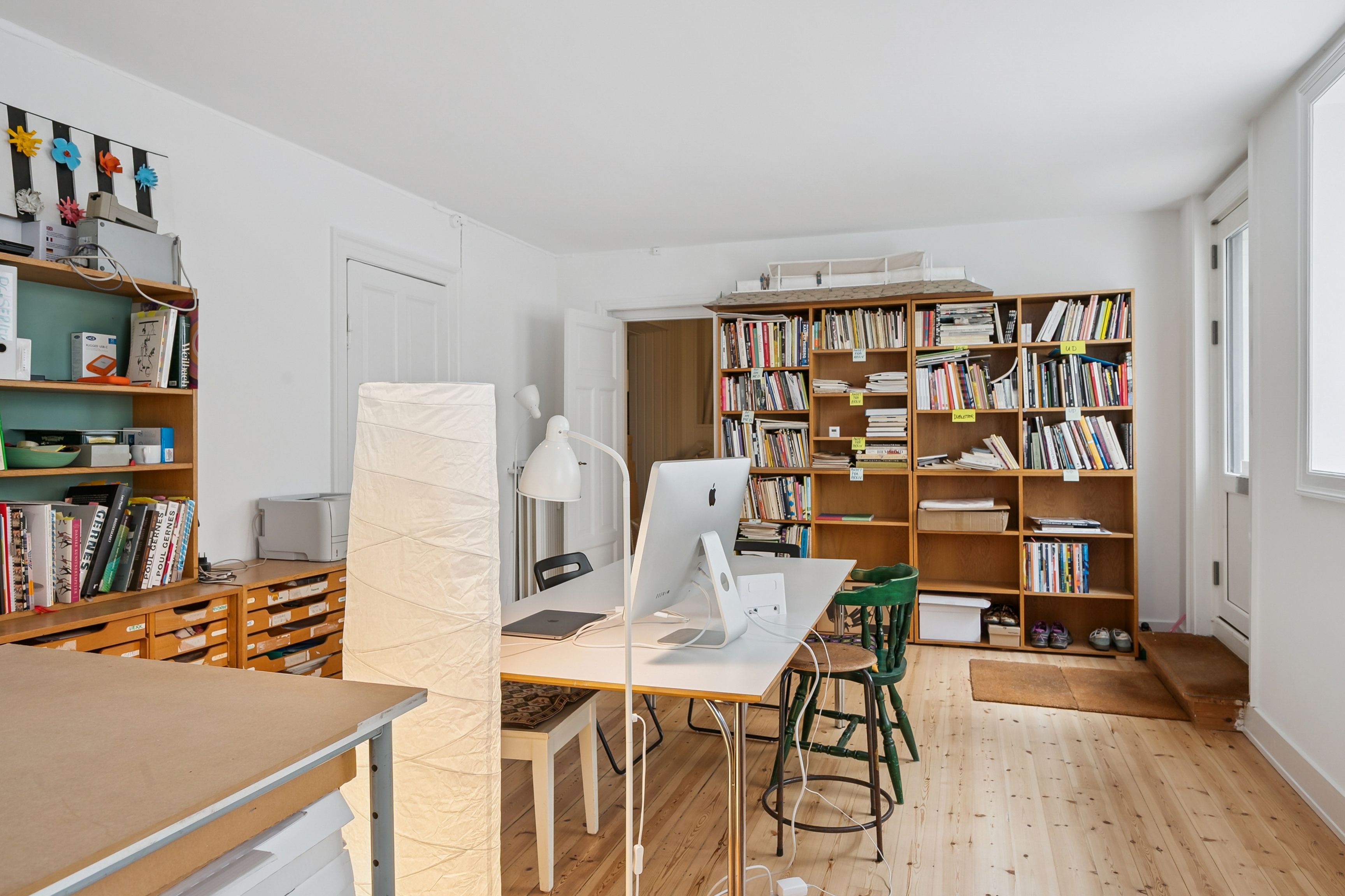 Et lyst kælderlokale med vinduer mod Collinsgade. Værelset rummer en bord med en computer, en lampeskærm og to stole. På