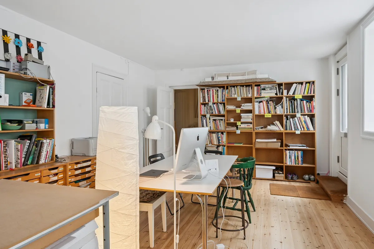 Et lyst kælderlokale med vinduer mod Collinsgade. Værelset rummer en bord med en computer, en lampeskærm og to stole. På