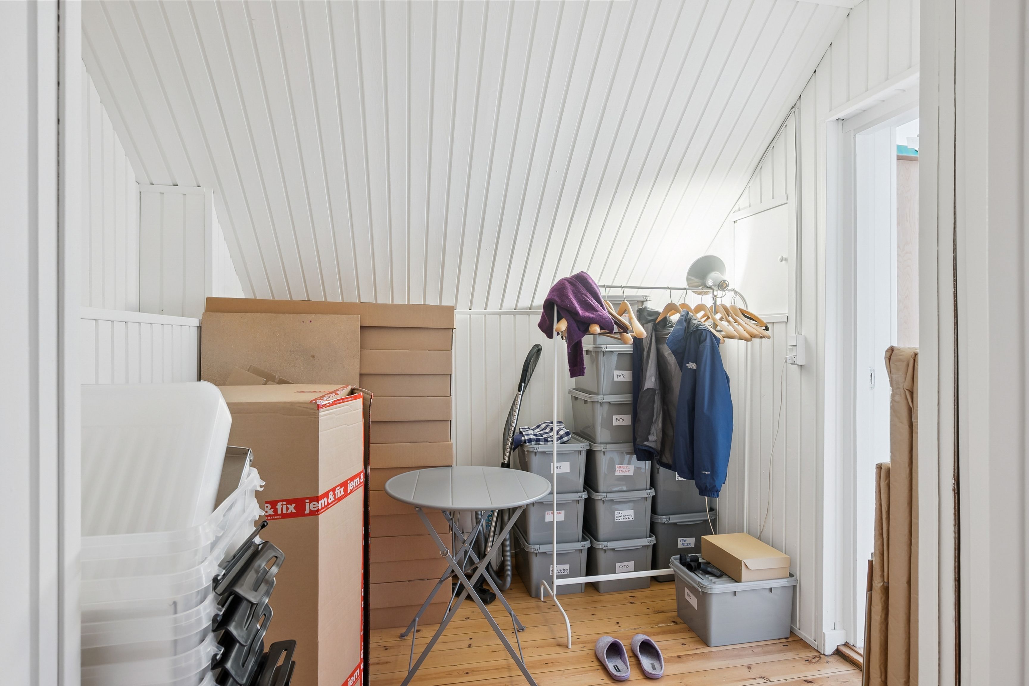 Et lille, lyst lager med flere kartoner og plastikkasser samt to par fodtøj og en lille rundt bord.