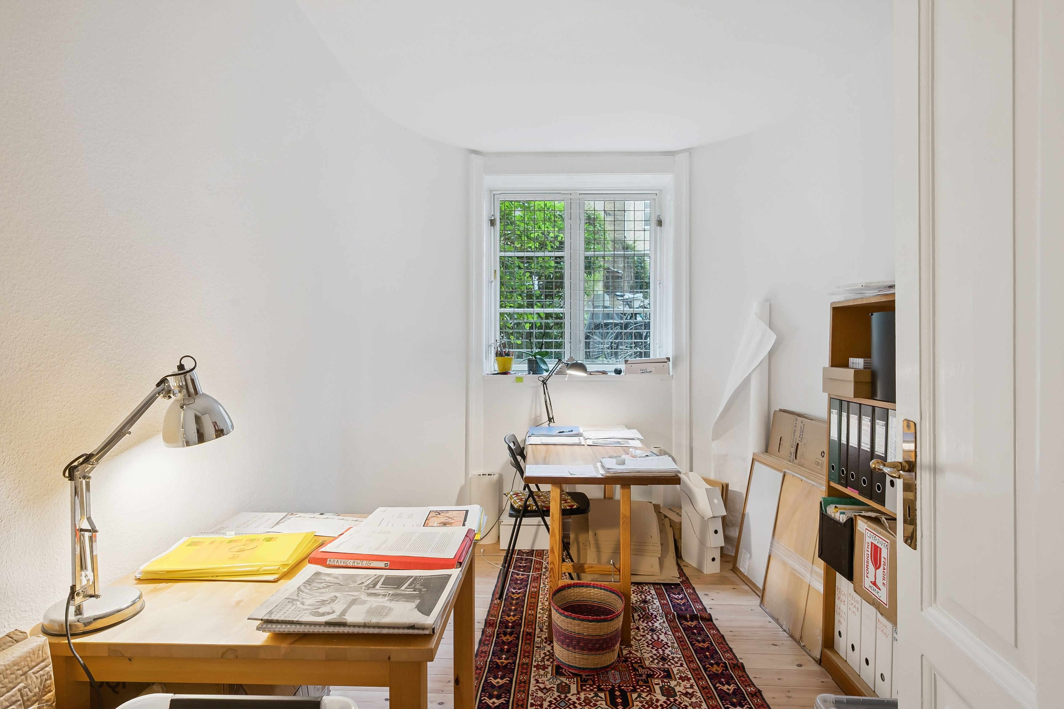 En lille arbejdsstue med to borde og en lampestang, der er placeret i en hjørne. Der er flere mapper og papirer på bordene,