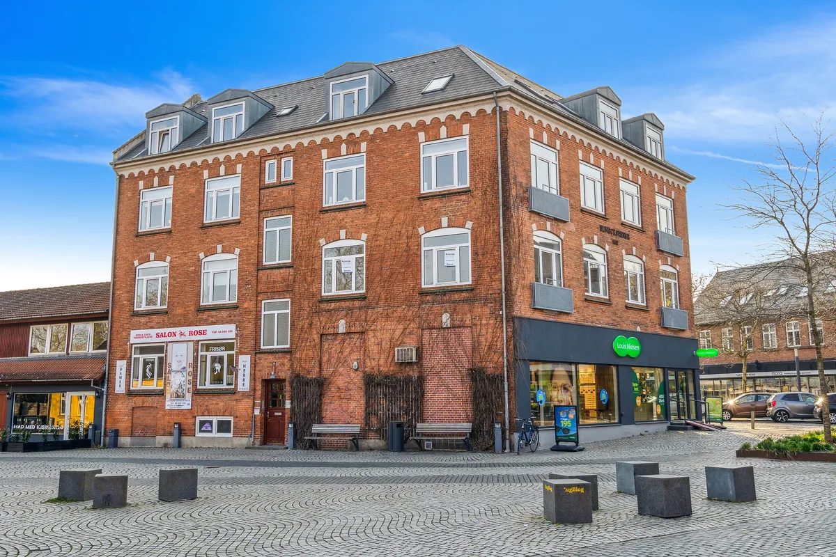 En stor murstensbygning med flere butikker og kontorer og flere bænke foran bygningen med en cykel og nogle planter og biler